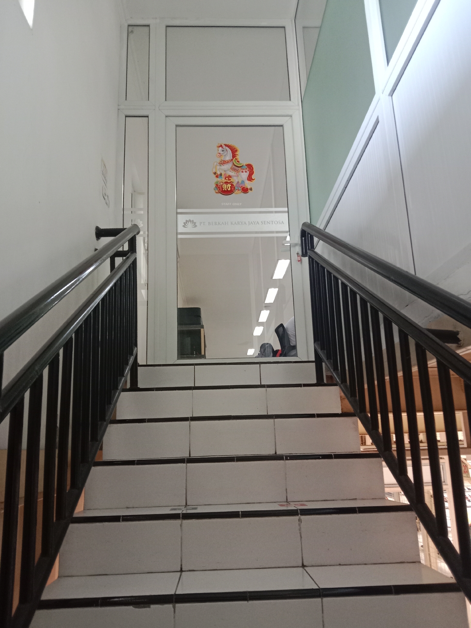 The image shows an interior view of a staircase with white tiled steps and black metal railings. The staircase leads up to a glass door with a decorative image of a unicorn and text that reads "STAFF ONLY" and "PT. BERKAH JAYA SENTOSA". The overall environment appears to be an office building, possibly in Semarang, Indonesia, given the company name. Natural light filters in from above, illuminating the stairway. There are no people clearly visible in the image.
