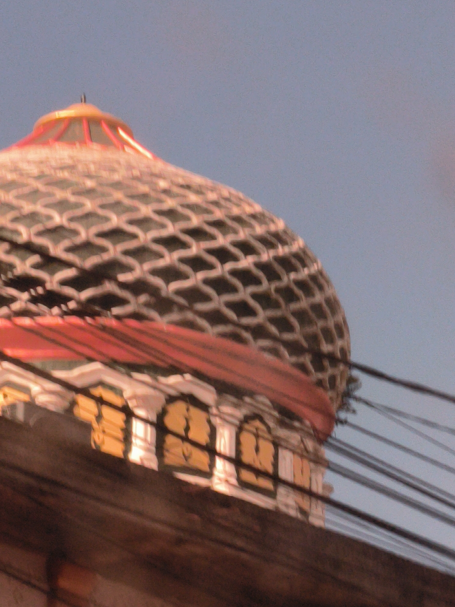 The image shows the upper portion of a building dome with a latticed pattern. The dome is decorated with a red band. The building has white and gold accents below the dome. Several power lines cross the foreground. The location is Idi Rayeuk, Indonesia.