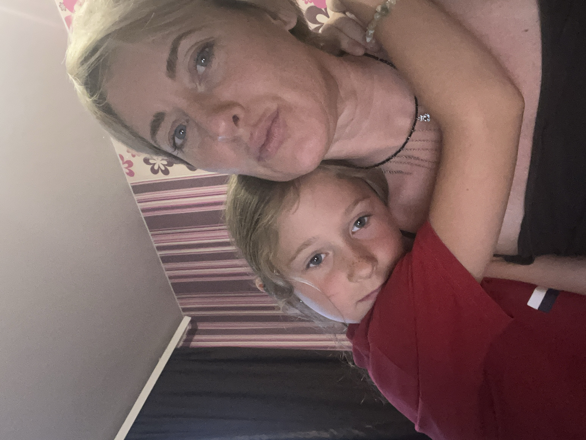A young child is being held close by an adult woman. The child is wearing a red shirt and is resting their head on the woman's shoulder, looking towards the camera with a slightly sad expression. The woman is looking towards the camera with a neutral expression. They are indoors, with a wall featuring striped wallpaper and dark curtains visible in the background. There is a ceiling visible at the top of the image. The scene appears to be a personal, informal moment captured in a domestic setting. There is no information in the image to suggest a location in the city of Saratov, Russia.