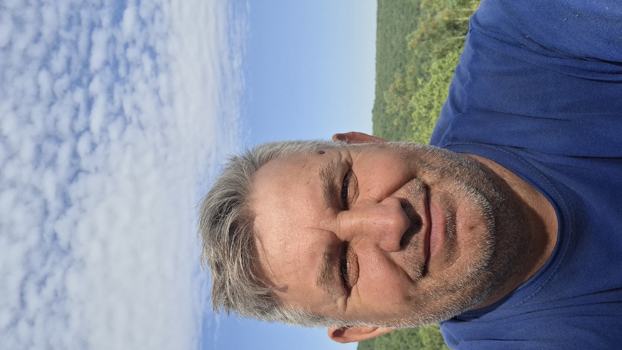 A man with graying hair and a beard, wearing a blue t-shirt, smiles into the camera. He has a mole on his left temple. The background shows a lush green forest against a bright blue sky with scattered white clouds. The image appears to be taken outdoors, likely in a rural or natural setting. There is no indication of the city of Hîncești, Moldova, within the image itself.