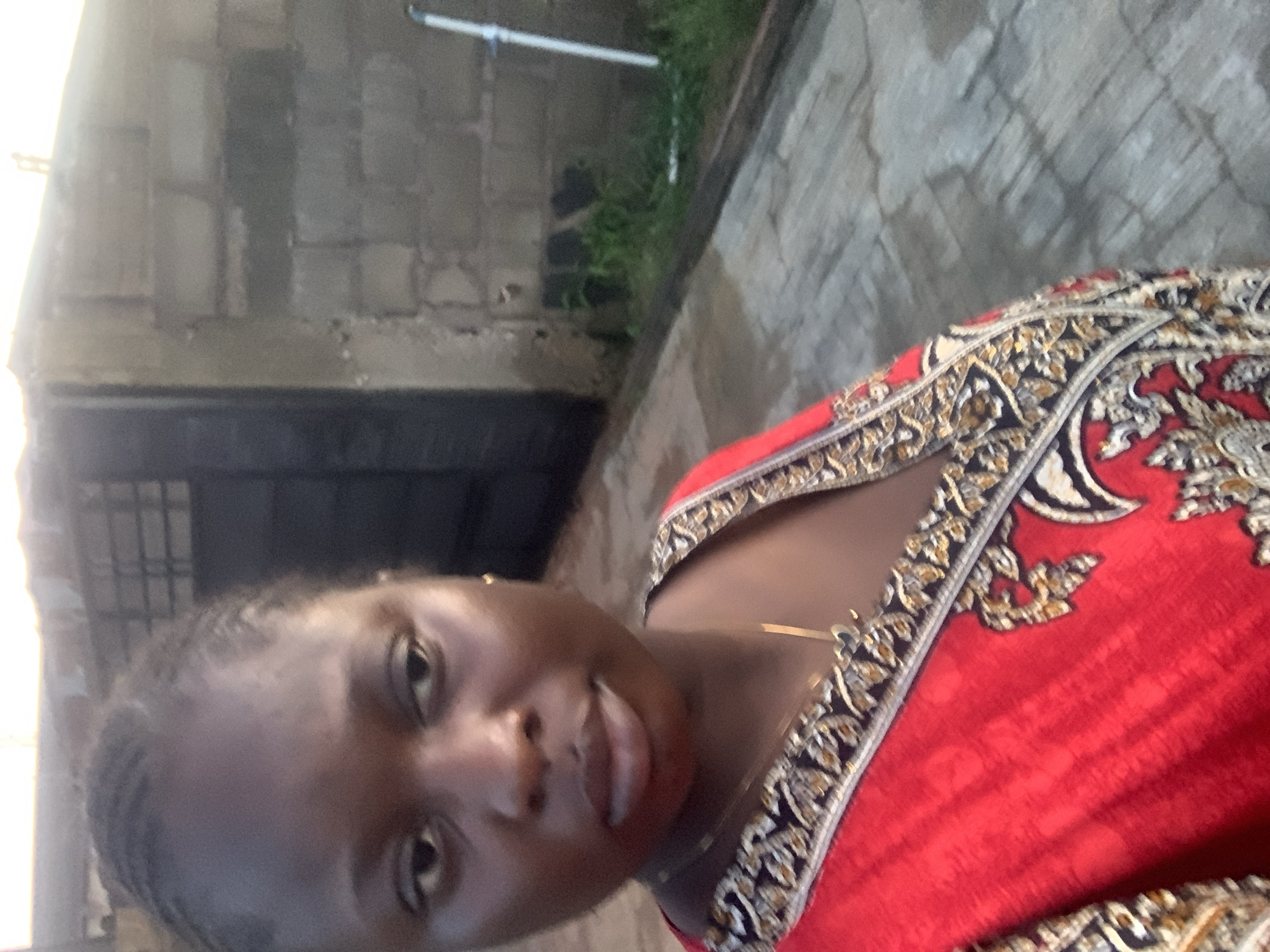 A close-up selfie shows a young Black woman from the chest up, looking slightly off-camera. She has braided hair and is wearing a red garment with an ornate black, gold, and white patterned neckline and a delicate gold necklace. The background is blurred and appears to be outdoors, with a rough brick wall to her right and a paved or concrete walkway in the foreground. The lighting suggests it is either early morning or late afternoon.