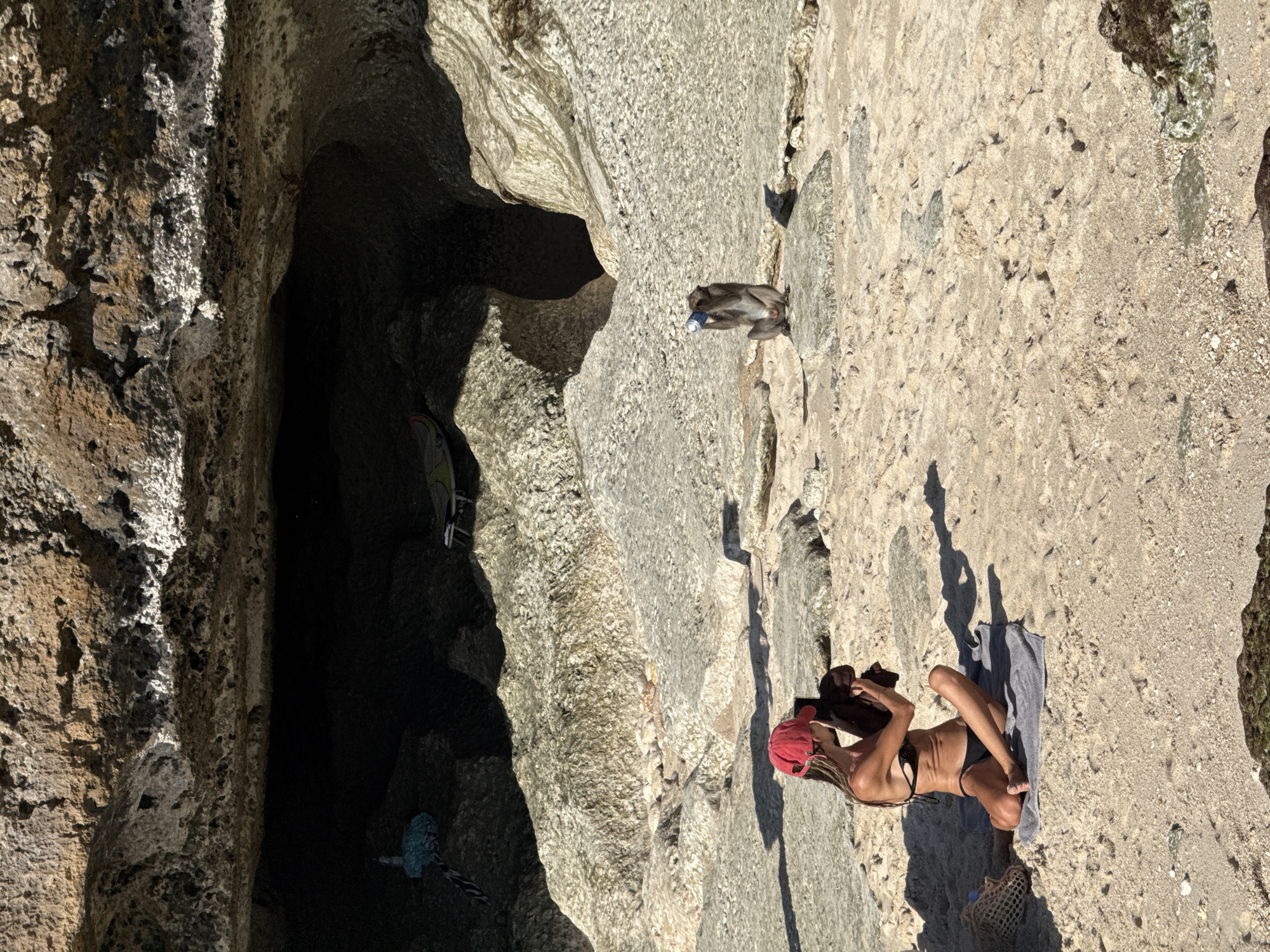 This image depicts a sunny scene at a rocky cove or beach in Kuta Selatan, Indonesia. In the foreground, a woman is seen relaxing on a grey towel laid out on light-colored sand and rocks. She is wearing a black bikini and a red baseball cap, and her hair, styled in dreadlocks, is visible beneath the cap. She appears to be looking down at a small object in her hands, possibly a phone. A mesh bag and a dark piece of fabric are lying near her.
Further into the scene, towards the left, a monkey is perched on a large, light grey rock. The monkey is holding a blue and white can, possibly a beverage. To the left, a prominent dark cave entrance is visible, characterized by rugged, textured rock walls. The interior of the cave is deep and shadowy. Inside or near the cave's opening, a colorful object and a blue braided rope-like item can be vaguely seen.
The time of day appears to be mid-day, with bright sunlight casting strong shadows, indicating clear weather. The ground consists of a mix of sandy patches and varied rock formations. No discernible text is visible on any objects in the image.