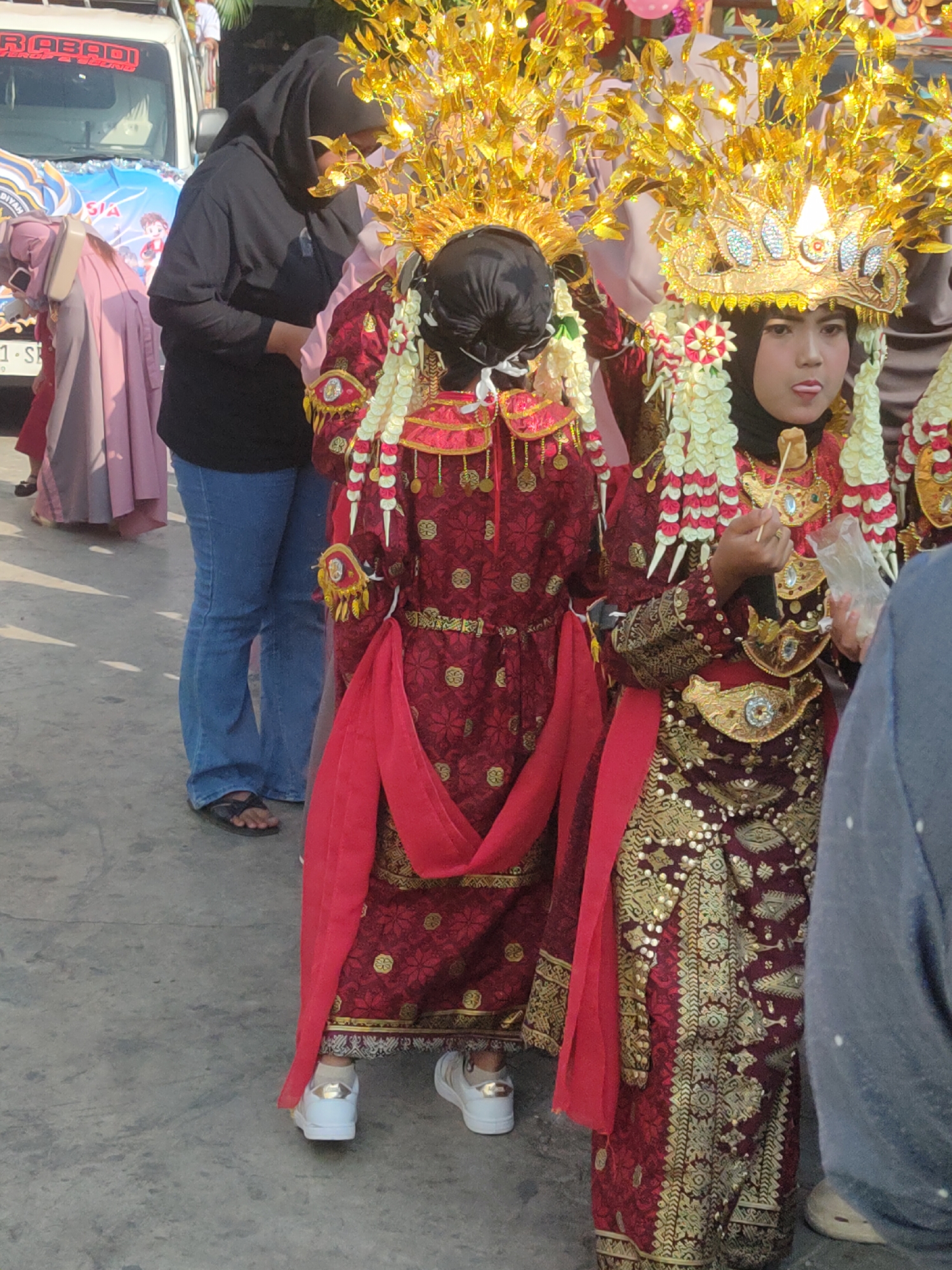 In the city of Payaman, Indonesia, two young girls are dressed in traditional attire for a celebration. They wear elaborate, ornate red and gold dresses and headdresses. The girl on the right has her tongue sticking out and holds a small piece of food on a stick. In the background, a woman in a black hijab and jeans stands near a white van decorated with colorful banners. Other people and a truck are also visible. The ground is paved and appears to be outdoors.