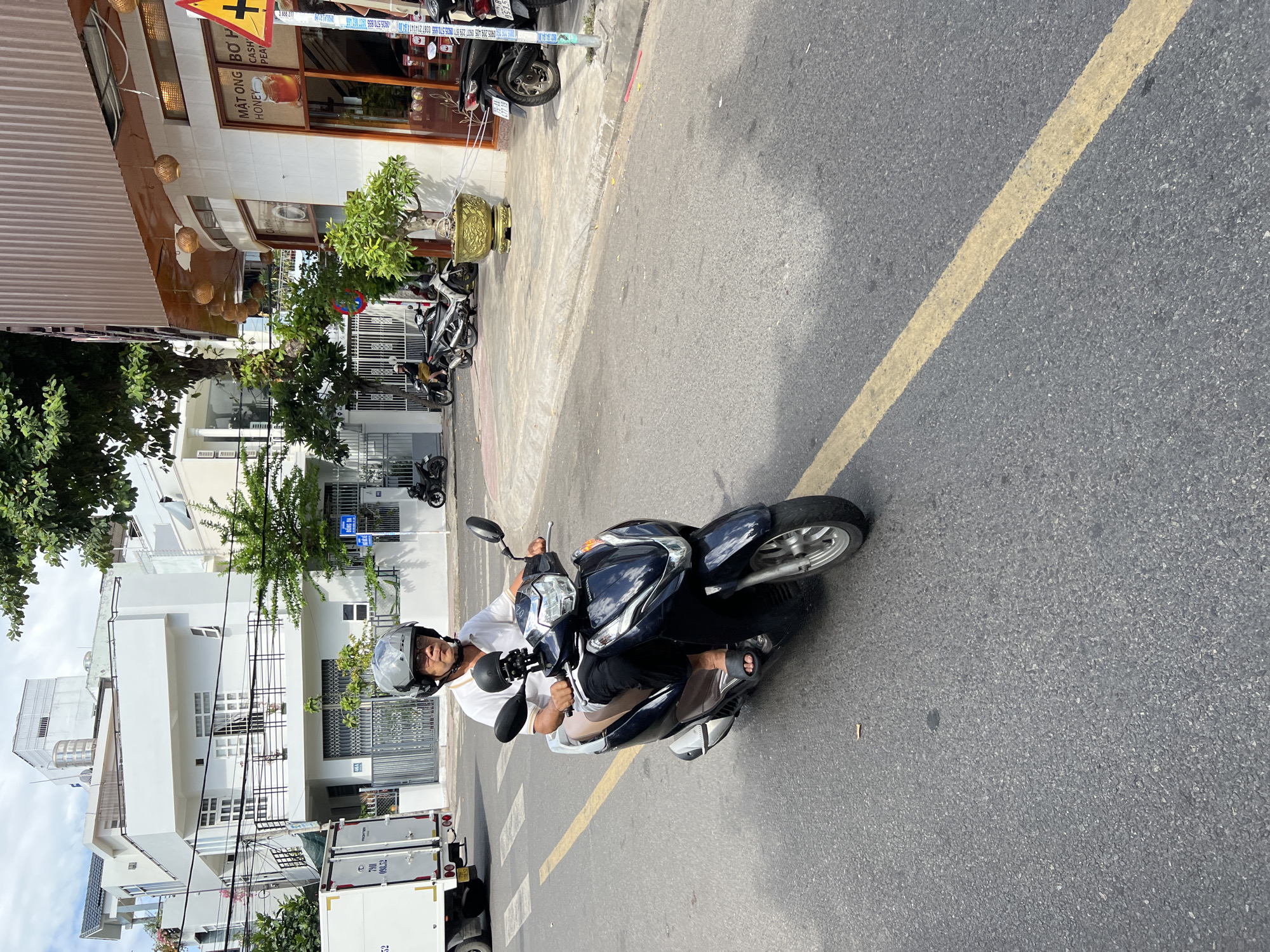 In Nha Trang, Vietnam, a man rides a dark-colored scooter on an asphalt road. He wears a white polo shirt, dark pants, and a grey helmet. The road has a yellow line down the center. On the right side of the road, there is a building with a brown metal facade and windows displaying signage for "MẬT ONG BƠ HẠNH PHÚC" and "HONEY CASH PEANUT". A yellow traffic sign with a black exclamation mark is visible. Several other scooters are parked on the sidewalk in front of the building. On the left side of the road, a white truck with the text "FRONTIER" is parked. In the background, several white buildings with balconies and trees are visible under a cloudy sky.