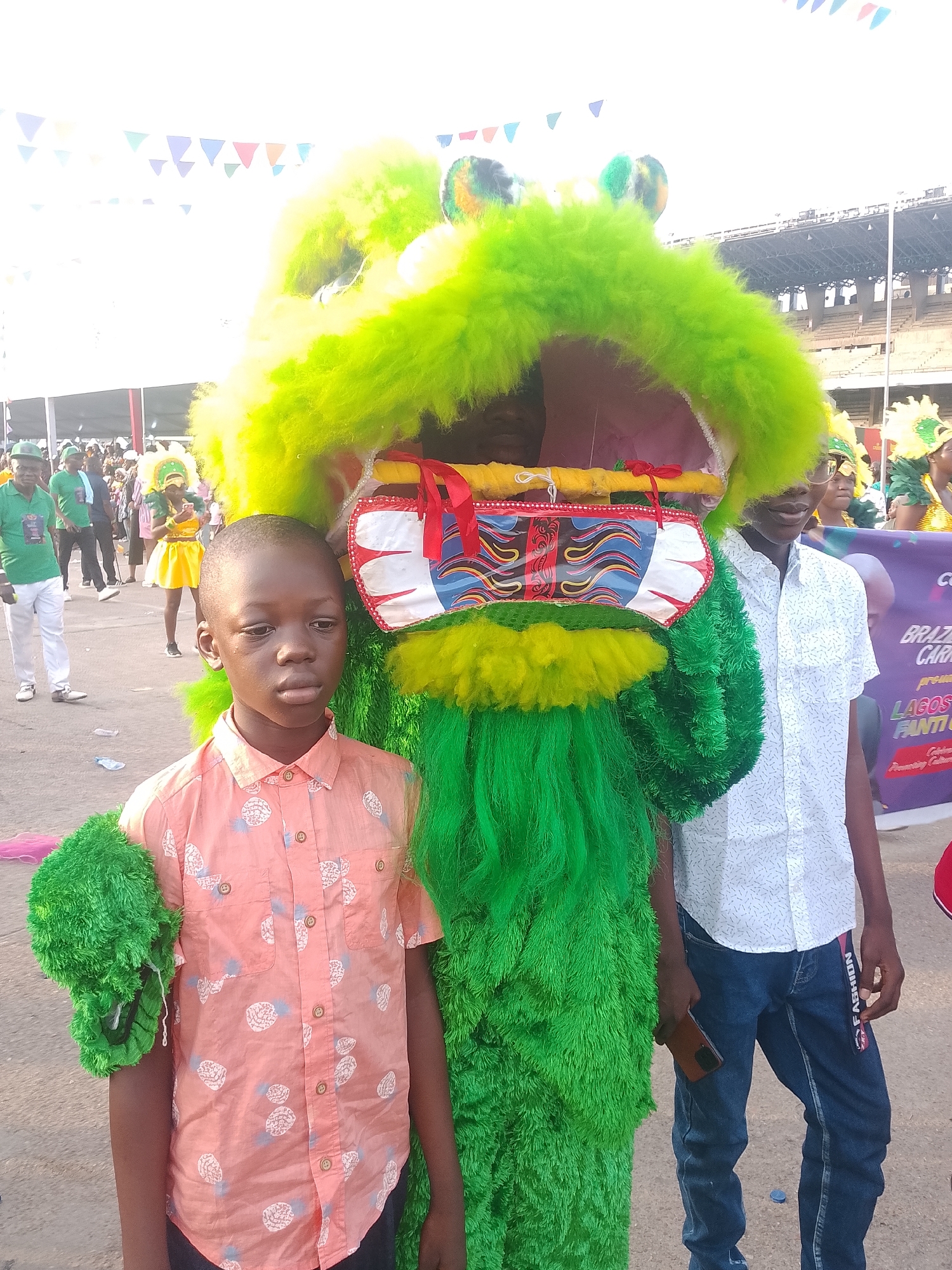 A medium-shot image depicts two boys standing next to a large, fuzzy, green costume, likely a dragon or similar creature, at a Lagos, Nigeria event. The younger boy, standing in front, wears a pink shirt with a pineapple pattern. He appears glum. The other boy is standing to the side and slightly behind the costume, he is wearing a white shirt with a small geometric pattern and jeans. The fuzzy green costume has a large open mouth, revealing inner decorations. In the background, crowds of people are visible, as well as colorful triangular pennants hanging above. A stadium is also visible in the background, suggesting an outdoor public event.