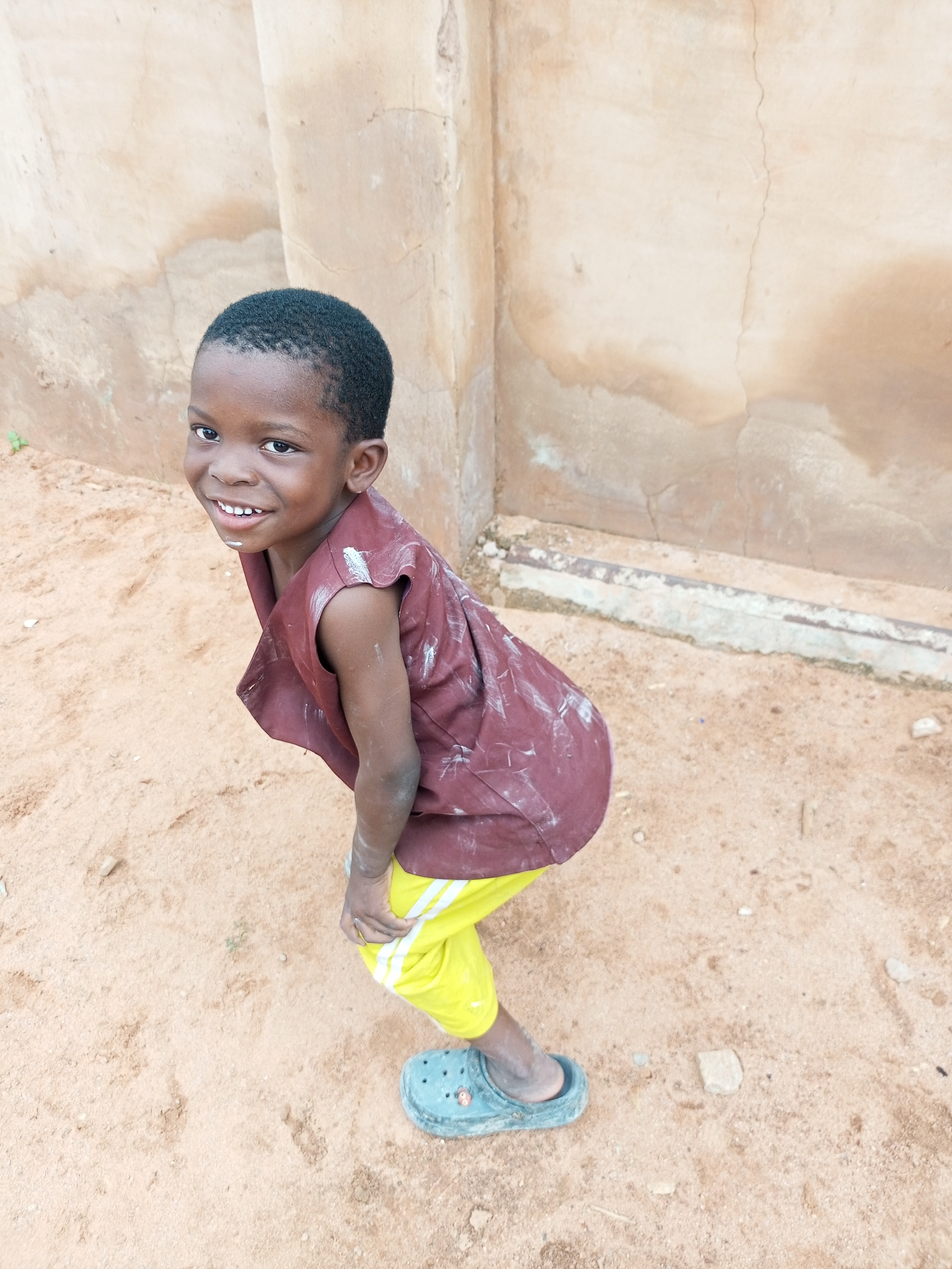 A young boy with dark skin and short black hair smiles at the camera while crouching in sandy soil. He wears a sleeveless, maroon tunic with white markings and bright yellow pants with white stripes. A single blue Croc sandal is on his left foot. The background consists of a rough, light-brown plastered wall with visible cracks and some staining. The ground is covered in fine sand with small stones and debris scattered about. A small green plant is visible to the left of the boy. The scene appears to be outdoors in a dry, possibly rural or semi-urban, environment in Tempure, Nigeria.