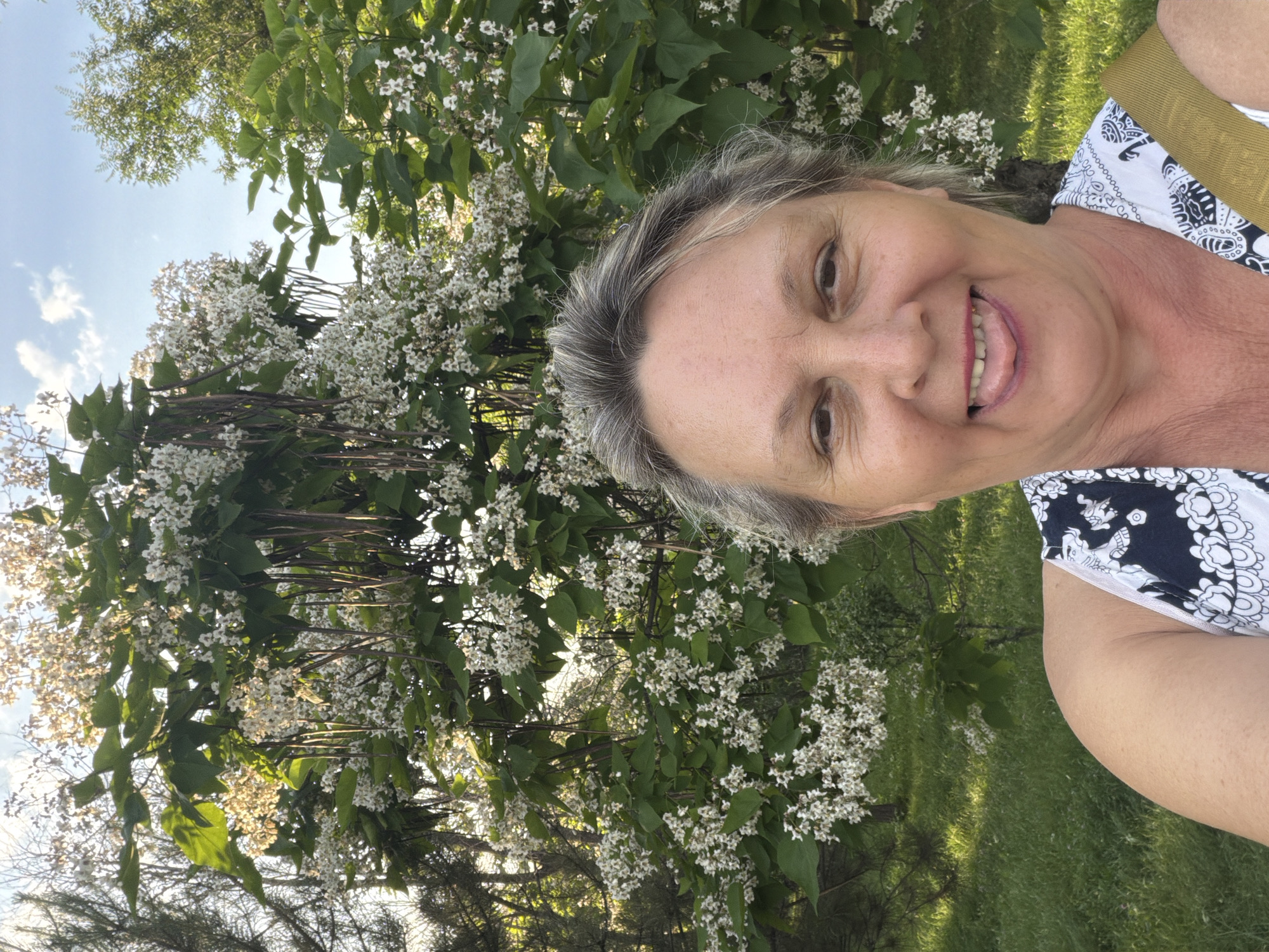 A woman with graying hair and fair skin smiles at the camera, sticking her tongue out slightly. She is wearing a white top with a black and white pattern. Behind her, a tree with numerous small white flowers and long seed pods is in bloom. The setting appears to be outdoors, with green grass and sunlight visible. The location is listed as Unknown, Unknown.