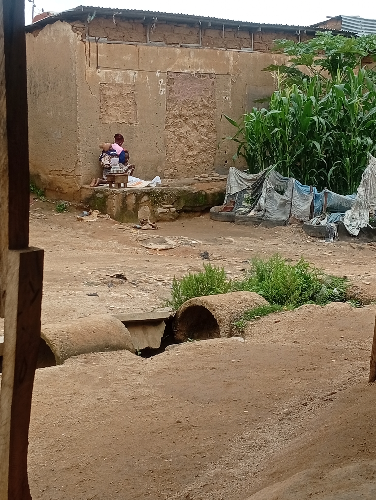The image depicts a street scene in Jos, Nigeria, characterized by a dirt road and rudimentary structures. In the foreground, a broken concrete culvert runs across the path, with some green vegetation growing around it. A person is seated on a low platform outside a building made of earth-colored material, with a corrugated metal roof. This person is holding a child who is wearing pink clothing. Next to the seated person is a small wooden stool with items on it. Behind the building, a dense patch of corn stalks is visible, along with some covered objects that appear to be tires wrapped in cloth. The overall impression is one of a rural or underdeveloped urban environment.