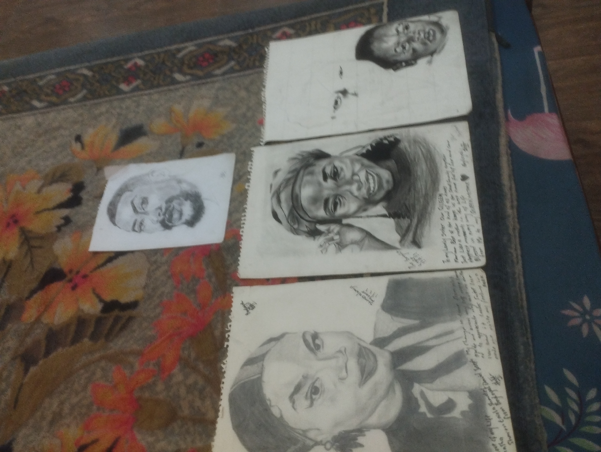 The image depicts several charcoal sketches of faces, laid out on a patterned rug. One sketch, at the top right, appears to be of a baby. Another, directly below it, shows a person with a wide smile and dark hair, possibly wearing a headwrap. To the left of these, there are two more sketches. The upper left sketch shows a portion of a face with darker hair. The lower left sketch, partially visible, shows a face with facial hair. The sketches are arranged in a slightly overlapping manner on a rug with a floral pattern featuring orange, yellow, and red flowers. A portion of a blue rug with a pink floral design is visible on the right side of the image. The overall setting appears to be indoors, with the rug likely on the floor. The scene is not specific to any particular location in Jos, Nigeria.