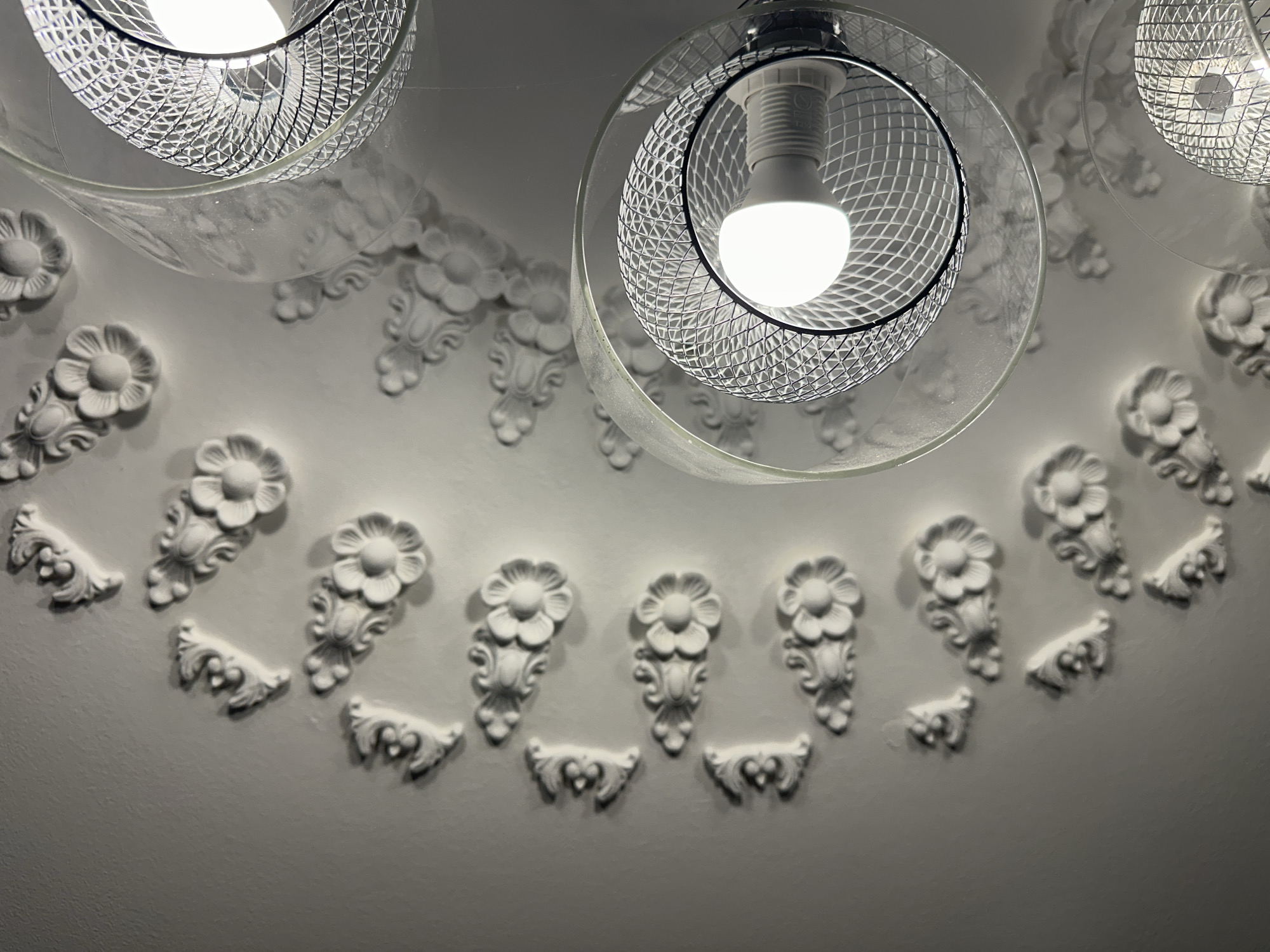 This image depicts an interior view of a ceiling with ornate plasterwork. The plasterwork features a repeating pattern of stylized floral motifs and scroll-like elements, arranged in a curved line. Two modern pendant lights hang from the ceiling. Each light fixture has a clear glass shade and a metallic mesh interior surrounding a lit bulb. The lighting creates distinct shadows of the mesh and the plasterwork on the ceiling. The image appears to be taken from a low angle, looking upwards. There are no people or animals visible in the image. The scene does not provide any specific contextual clues that would allow for the identification of its location within the city of Yessentuki, Russia.