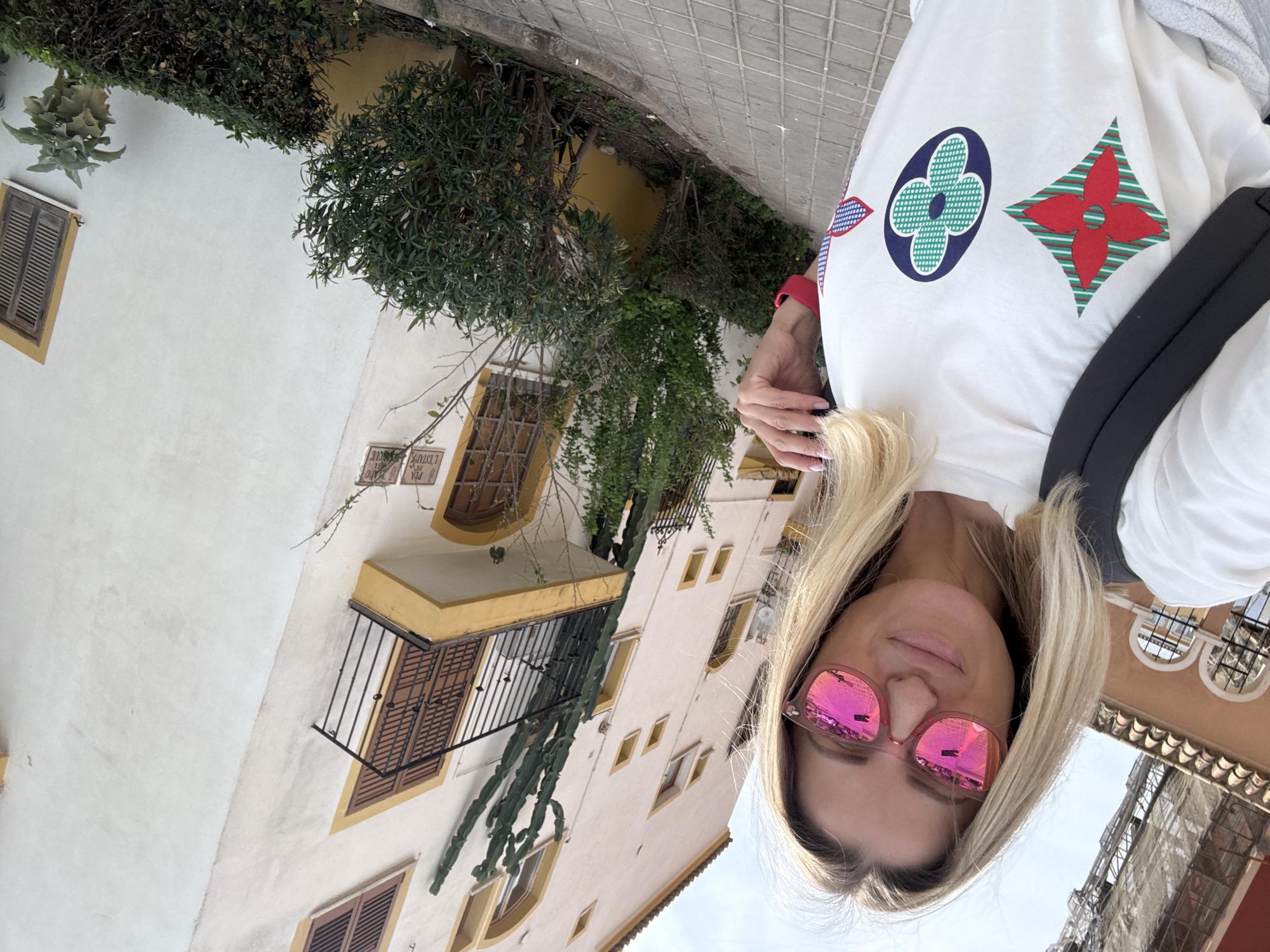A selfie taken by a blonde woman wearing pink sunglasses and a white t-shirt with large geometric patterns in a street with buildings. The buildings are mostly white with yellow trim around the windows and balconies. Some have brown wooden shutters. Plants and greenery can be seen growing on and around the buildings. The setting is in Urbanització Port Saplatja, Spain.