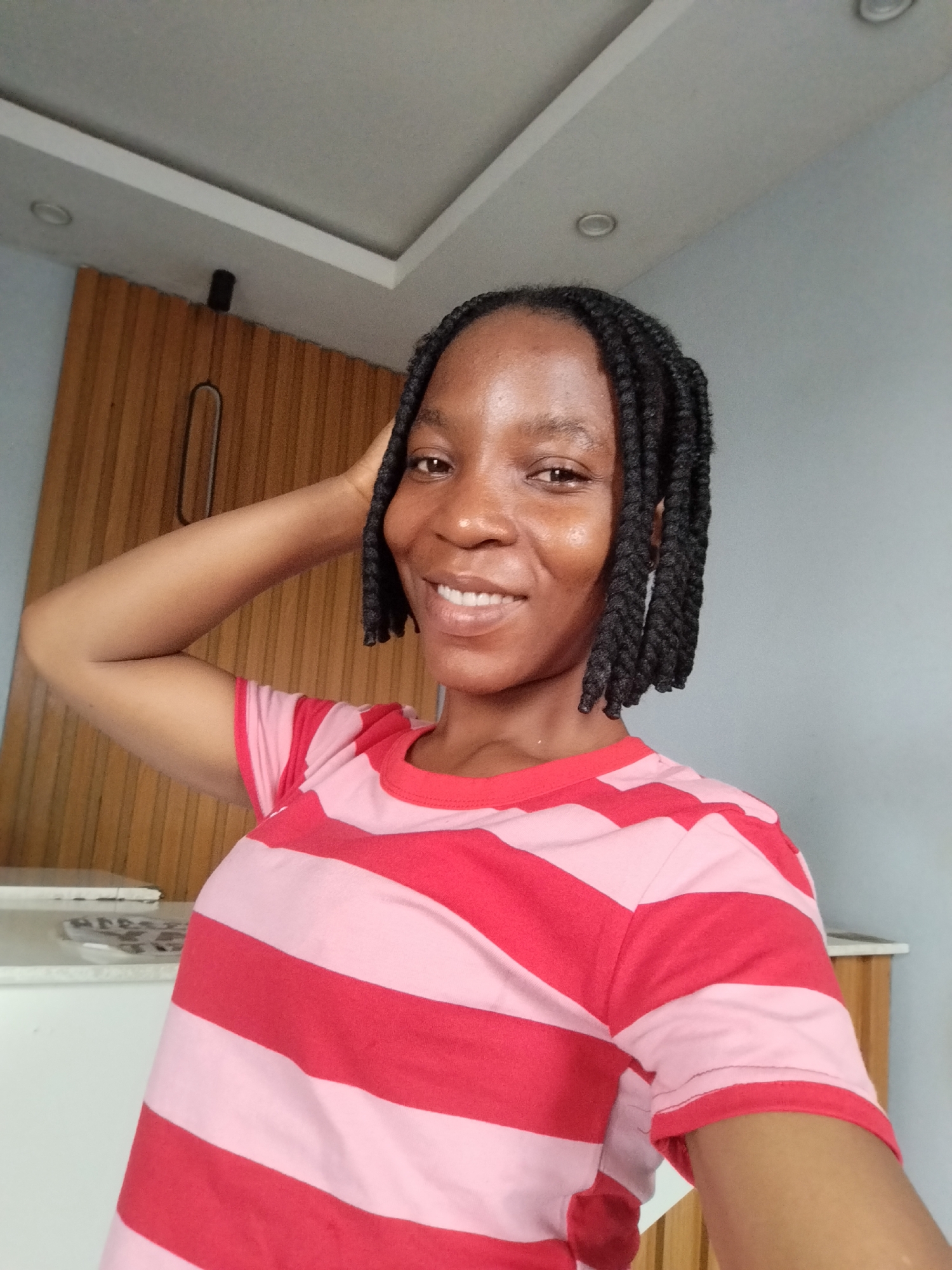 A woman with braided hair is in a room. She is smiling and has her right hand on her head. She is wearing a red and pink striped t-shirt. The background is a wall with wooden accents and a ceiling with lights. The image is captured from a selfie perspective. The location is Uselu, Nigeria.
