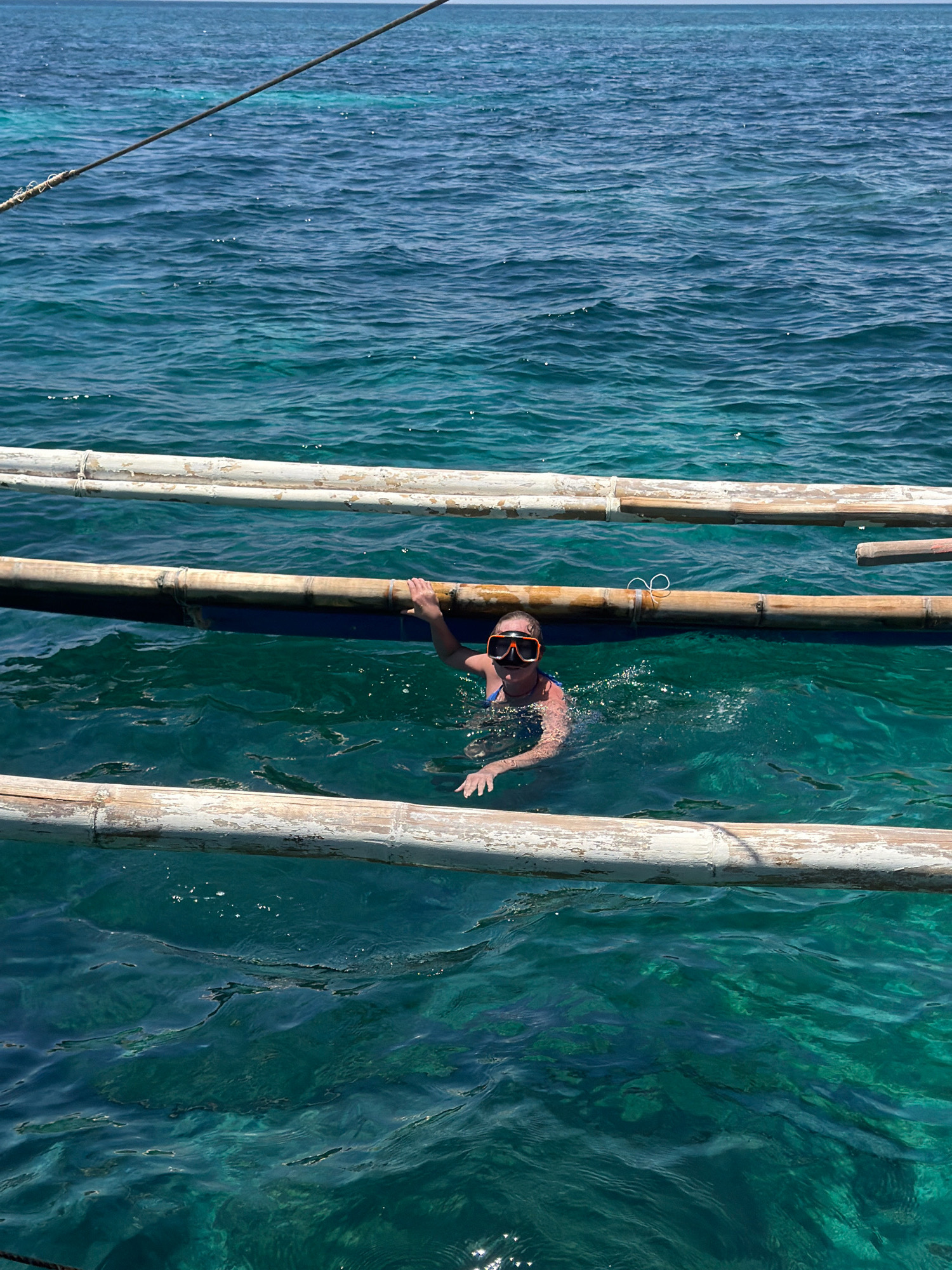 A woman wearing a blue swimsuit and orange snorkel mask is swimming in clear turquoise water near a wooden outrigger boat in Malay, Philippines. She is partially submerged, with her head and shoulders visible above the water. Her left hand is resting on the bamboo outrigger of the boat, and her right hand is submerged in the water. The water is clear, allowing a view of the sandy bottom beneath. The sun is shining, casting dappled light on the water's surface. The scene appears to be a tourist activity, likely snorkeling or swimming from a boat.
