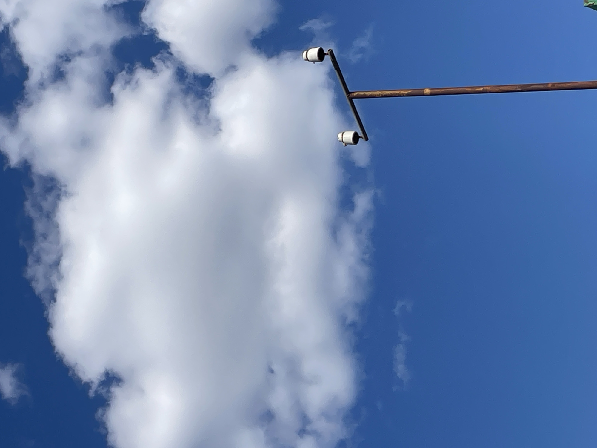 A rustic brown metal pole, weathered and aged, rises vertically against a vibrant, clear blue sky. At the top of the pole, a horizontal crossbar extends outwards, supporting two cylindrical white insulators. Large, fluffy white clouds dominate the upper portion of the frame, their soft edges contrasting with the sharp blue of the sky. The scene appears to be an outdoor setting, likely in Rostov-on-Don, Russia, given the context. No people or specific actions are depicted.