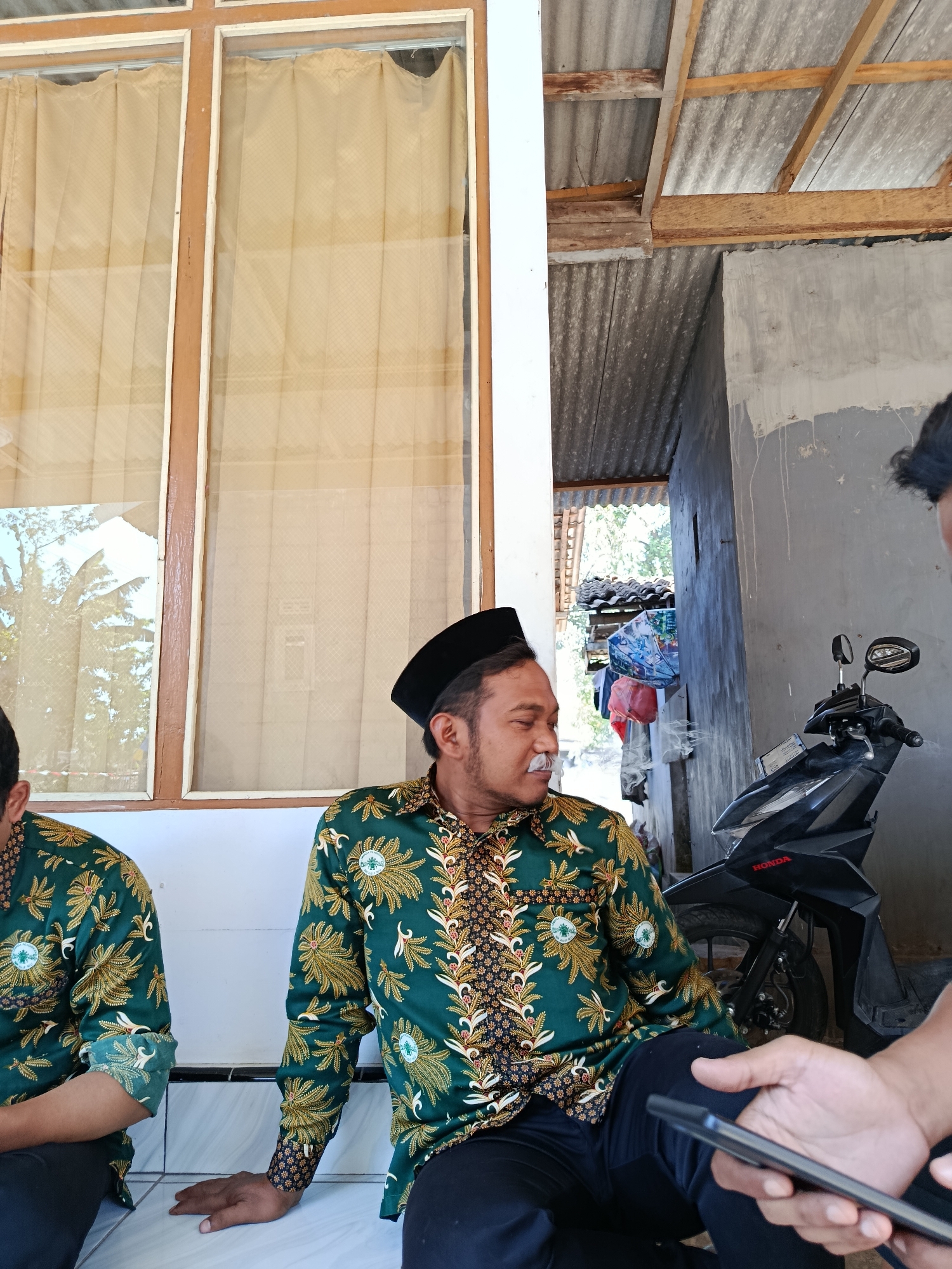 In Sempol, Indonesia, two men, likely in their 30s or 40s, are seated on a white tiled ledge outdoors. The man on the right, facing left and slightly away from the camera, wears a black peci cap and a green batik shirt with a gold floral pattern. He has a mustache and is exhaling smoke, suggesting he is smoking. The man on the left is partially visible, also wearing a similar green batik shirt. In the background, a black Honda scooter is parked. The environment appears to be a simple outdoor area with a corrugated metal roof supported by wooden beams, and a wall made of rough concrete. The area also features a window with tan curtains.