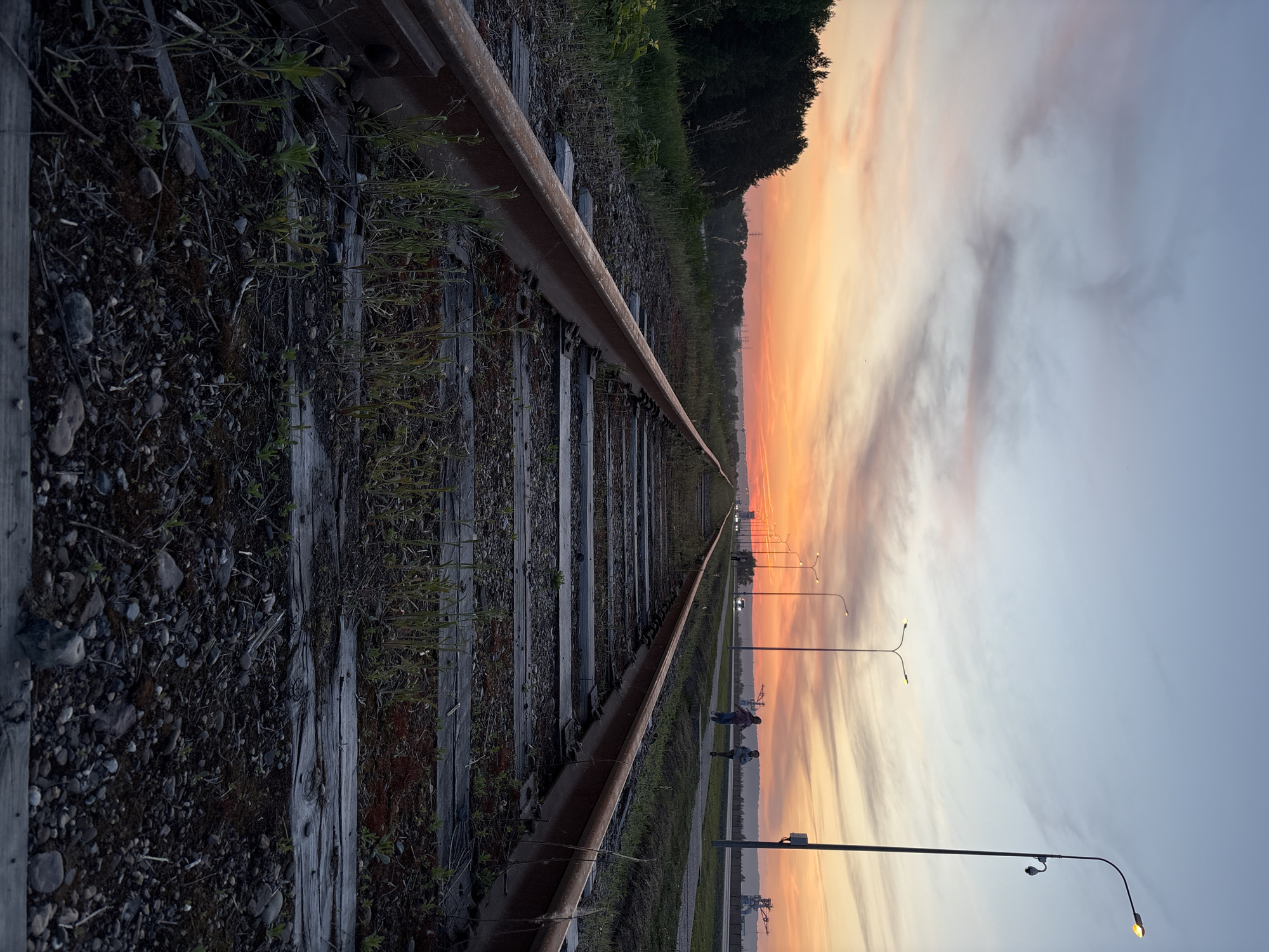 The image shows a railway track in Chaikovsky, Russia, receding into the distance at sunset. The tracks are flanked by ballast and vegetation. A road with streetlights runs parallel to the tracks. Two figures are visible walking beside the tracks near the road. The sky is a mix of orange, yellow, and grey hues. Cranes are visible in the distance.