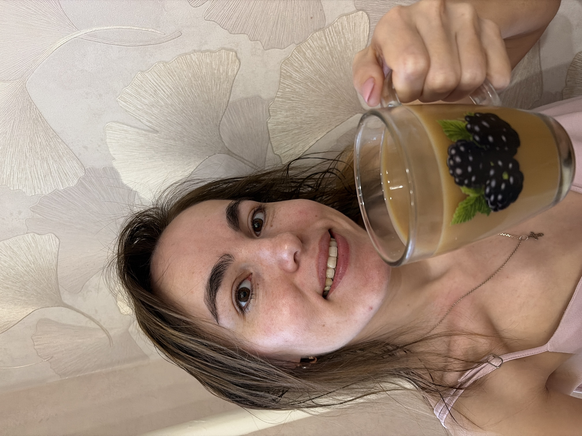 A woman with brown hair is holding a clear mug filled with a tan liquid, possibly coffee or tea with milk. The mug has a design of blackberries on it. The woman is smiling and looking at the camera. She is wearing a light pink top and a thin silver necklace with a small cross pendant. The background is a light-colored wall with a pattern resembling stylized ginkgo leaves. There is no explicit indication that the location is Izhevsk, Russia.