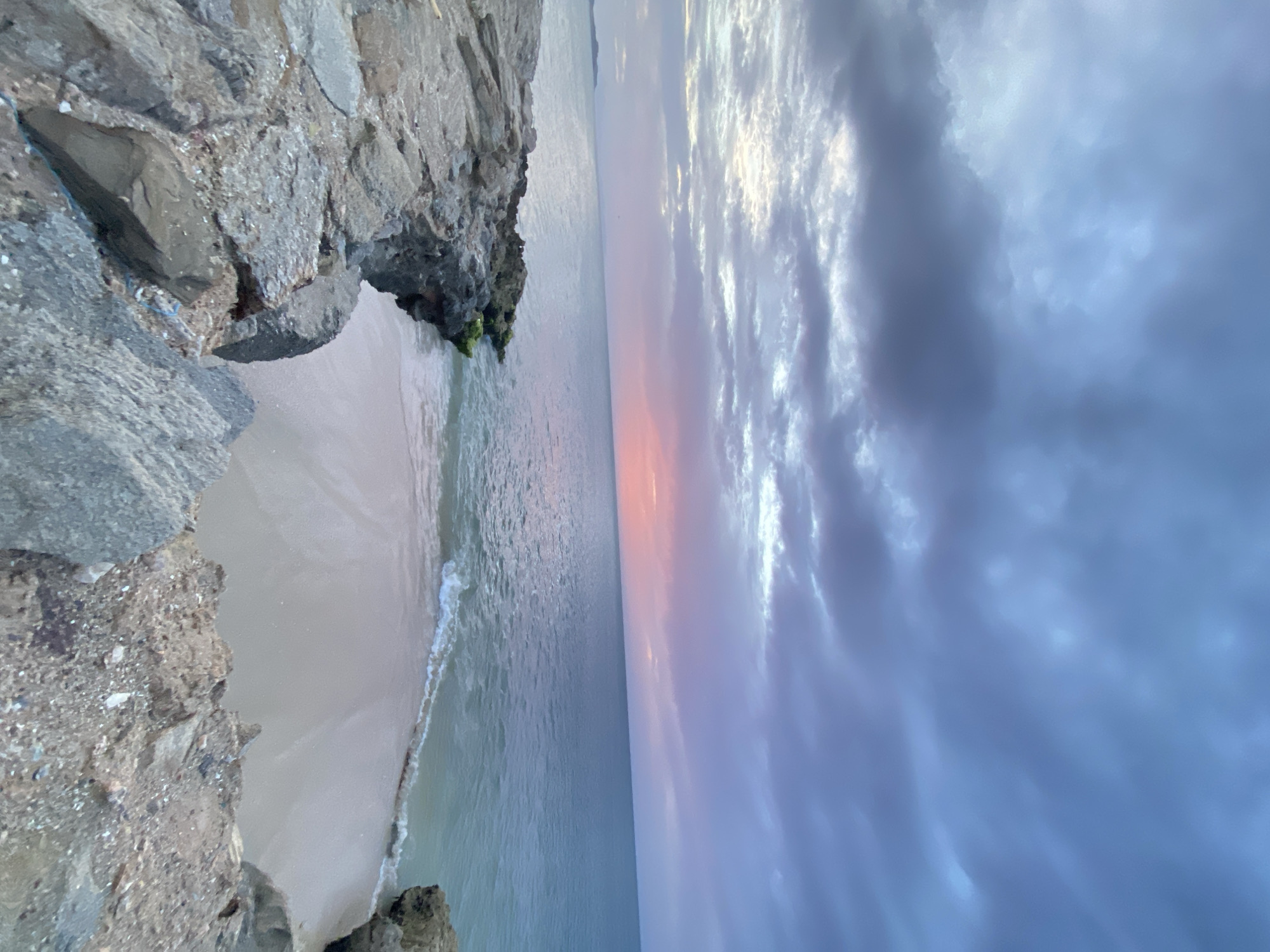 A wide-angle, low-angle shot captures a serene coastal scene at dusk in Juan Griego, Venezuela. The foreground is dominated by a rugged, rocky shoreline, with large, grey stones and some patches of moss. The rocks form a natural barrier that frames a small, sandy beach. Gentle waves lap at the shore, creating a soft white foam.
The sea stretches out to the horizon, a tranquil expanse of muted blue and green. The sky above is a dramatic canvas of clouds, painted with hues of grey, purple, and soft pink, suggesting the last moments of sunset. The light is diffused, creating a peaceful and slightly melancholic atmosphere. In the distance, a faint landmass is visible on the horizon. There are no people or animals present in the image. The overall impression is one of natural beauty and quietude.