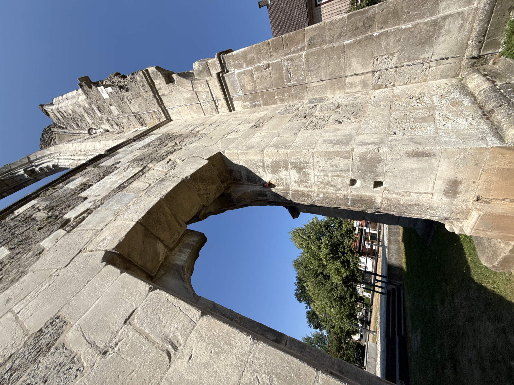 The image shows a close-up, low-angle shot of the ruins of the Walkenried Abbey in Walkenried, Germany. The dominant feature is a large stone archway and a portion of a weathered stone wall, made of large, rectangular blocks. The stonework shows signs of age and erosion, with some rough textures and discolored patches.
Through the archway, a clear blue sky is visible, along with a section of a grassy area with some trees and a paved pathway. Beyond the trees, a building with a brown tiled roof and light-colored walls can be seen. The sunlight casts shadows on the ruins and the ground, indicating a bright, sunny day. There are no people or animals visible in the image. The ruins appear to be part of a historical structure, likely an abbey given the location.