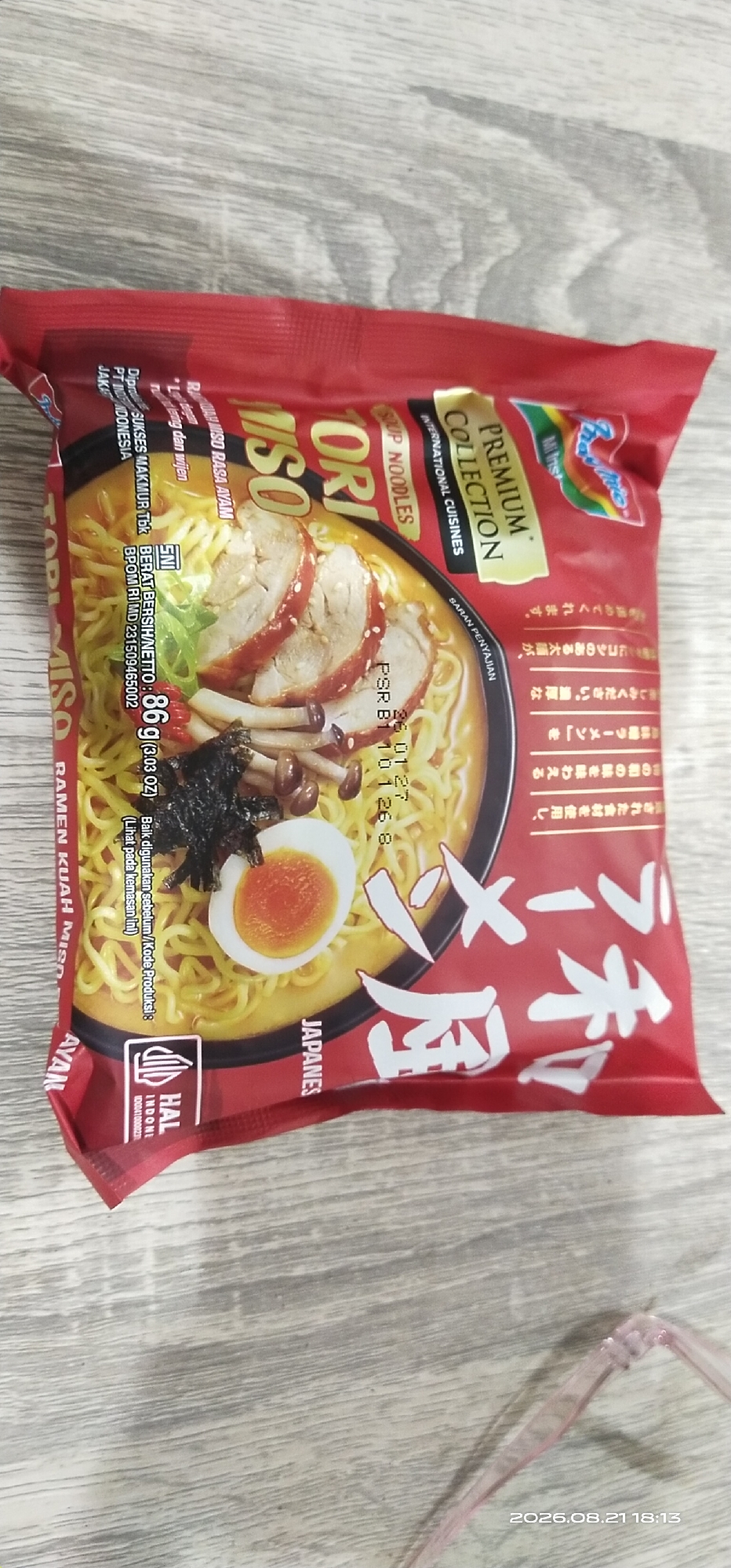 The image shows a package of Indomie Ramen Kuah Miso Ayam soup noodles on a wooden surface. The package is predominantly red with text and imagery of the soup, including noodles, slices of meat, mushrooms, and a boiled egg. There are also Indonesian and Japanese labels on the packaging. The bottom left corner of the package displays a HALAL certification logo. The image is a close-up, top-down view of the product. There is no indication of the city of Matabu, Indonesia within the image itself.