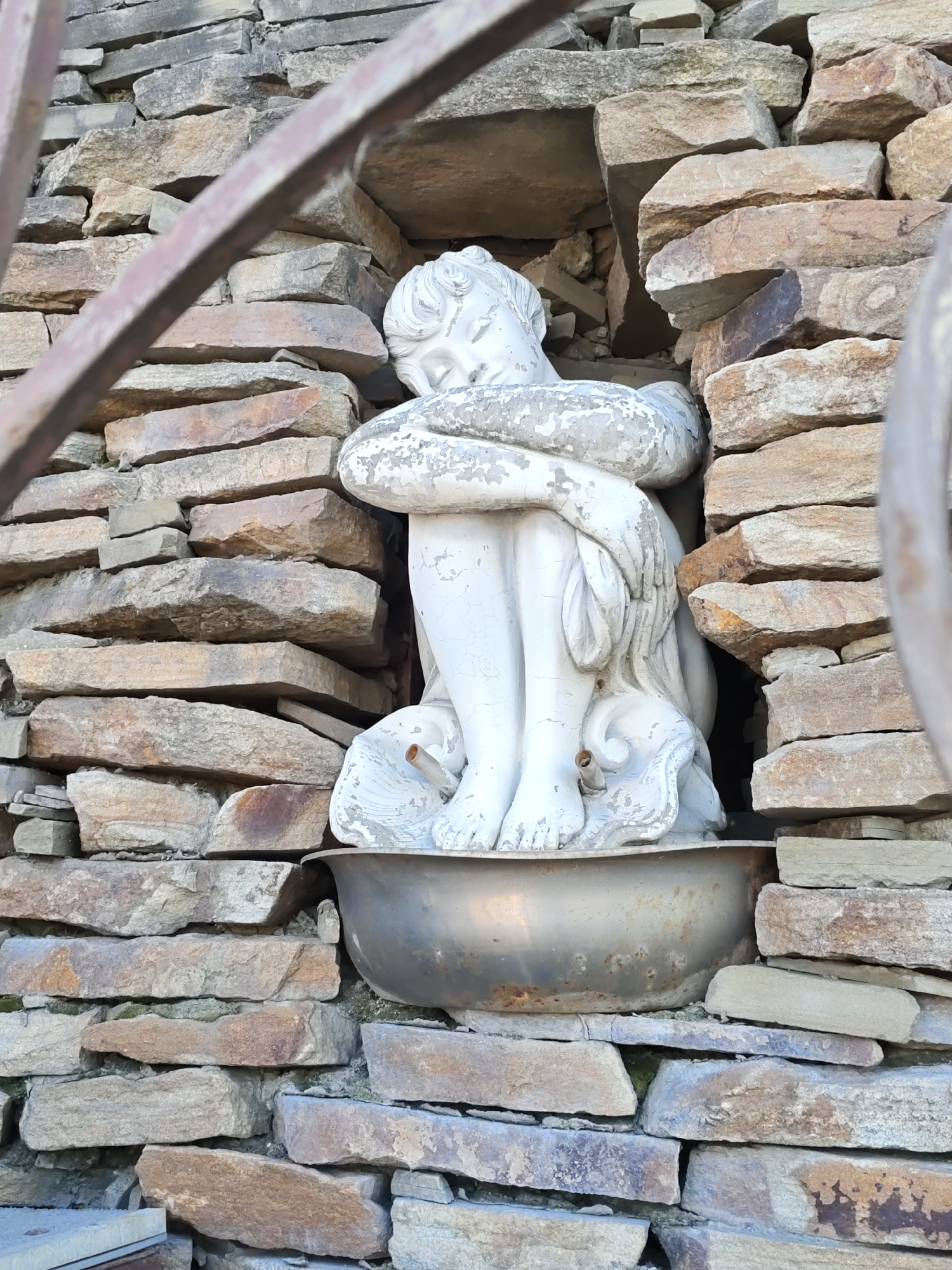 A white statue of a curled-up child sits within a niche in a stone wall. The statue is positioned in a metal basin. The stone wall is constructed from irregular, flat stones of varying sizes and colors, predominantly shades of beige and brown. The statue is partially obscured by a rusty metal railing in the foreground. The overall scene appears to be outdoors. The specific location in Rostov, Russia cannot be determined from the image alone.