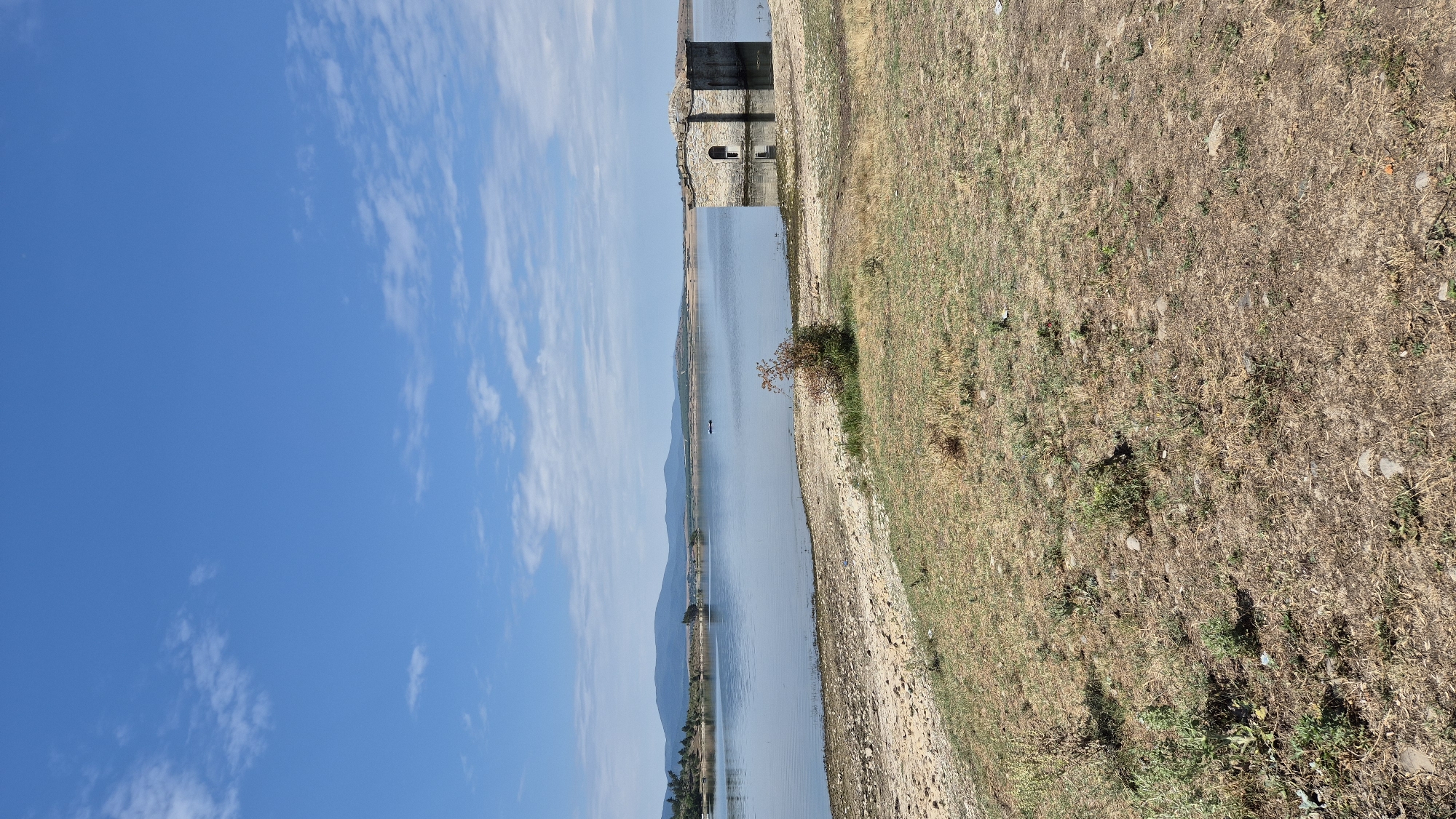 The image depicts a partially submerged stone structure in the middle of a calm body of water. The structure appears to be a building, possibly a church or chapel, with an arched doorway visible. The water level is low enough to reveal the base of the structure and a rocky shoreline. The surrounding landscape is dry and covered in sparse, yellowish-brown vegetation. In the distance, rolling hills form the horizon under a bright blue sky with scattered white clouds. Two small, dark objects, possibly boats or buoys, are visible on the water in the mid-ground. The scene is bathed in sunlight, suggesting a clear day. The location is identified as Panicherevo, Bulgaria, known for its reservoir which sometimes reveals submerged structures due to fluctuating water levels. There are no people visible in the image.