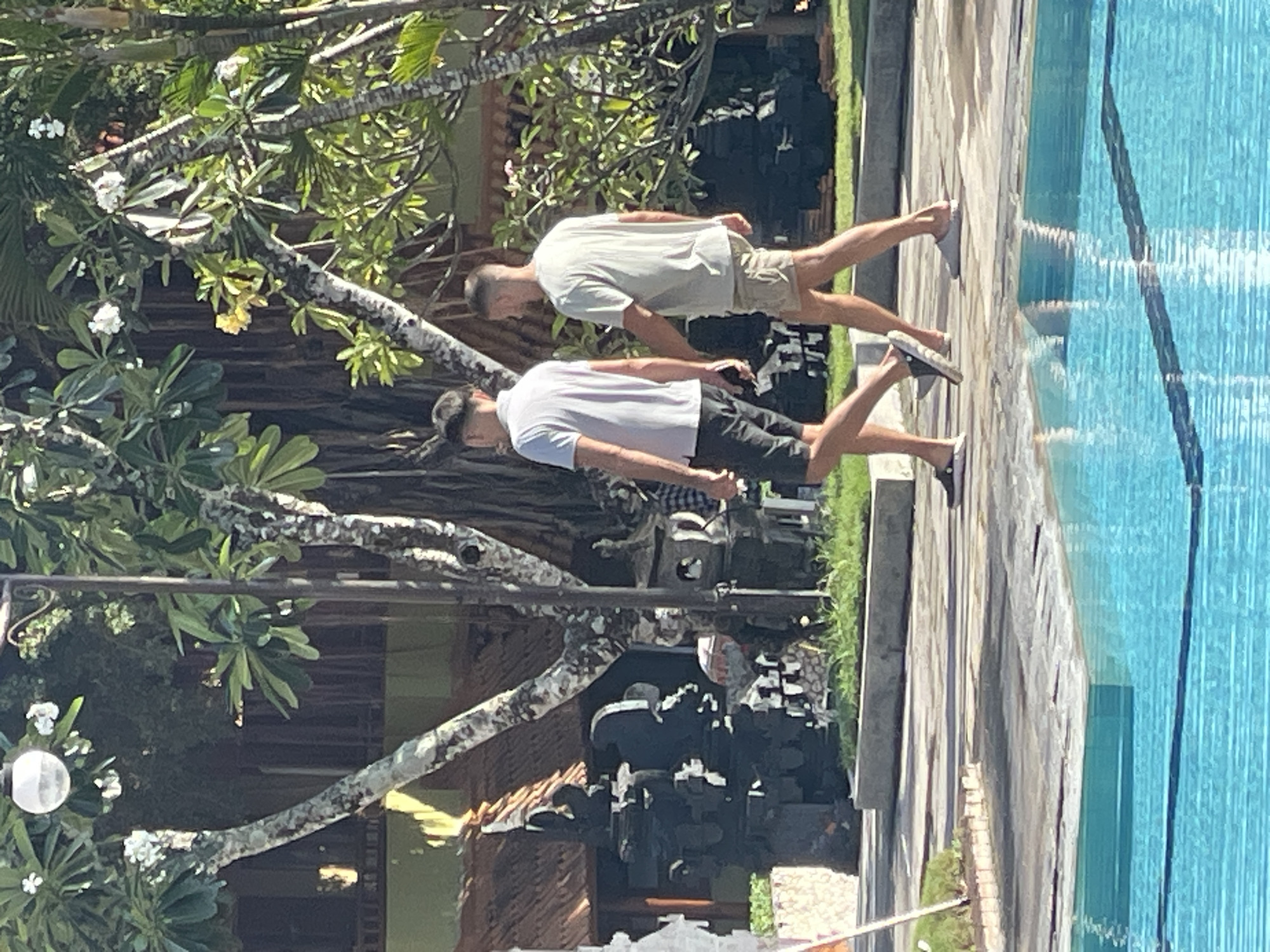 Two men are walking along a paved path next to a swimming pool. The man on the left is wearing a white t-shirt and black shorts. The man on the right is wearing a light-colored t-shirt and beige shorts. They are walking away from the camera, and their bodies are angled to the left. The path is lined with grass and trees, and in the background, there is a building with a thatched roof and some dark, stone statues. The swimming pool is filled with bright blue water. The scene appears to be a resort or hotel in a tropical location, likely Kuta Selatan, Indonesia, given the architectural style and lush vegetation.