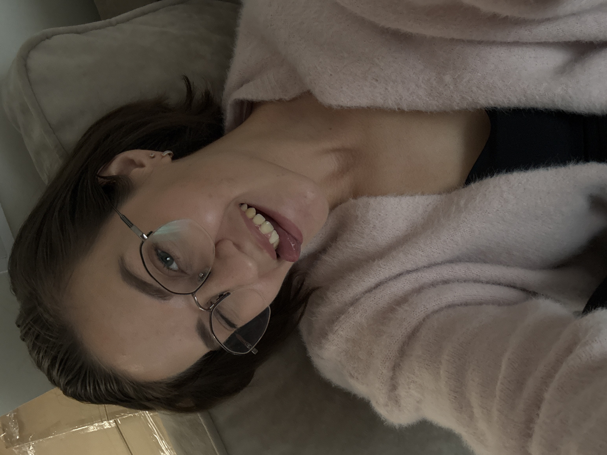 A young woman with brown hair and eyeglasses is lying on a gray upholstered surface, possibly a sofa or bed. She wears a light pink, fluffy sweater and a dark top underneath. She is sticking her tongue out towards the viewer. The background includes a wall and what appears to be a cardboard box. The location is not identifiable as Pervomaysky, Russia, from the given visual information.