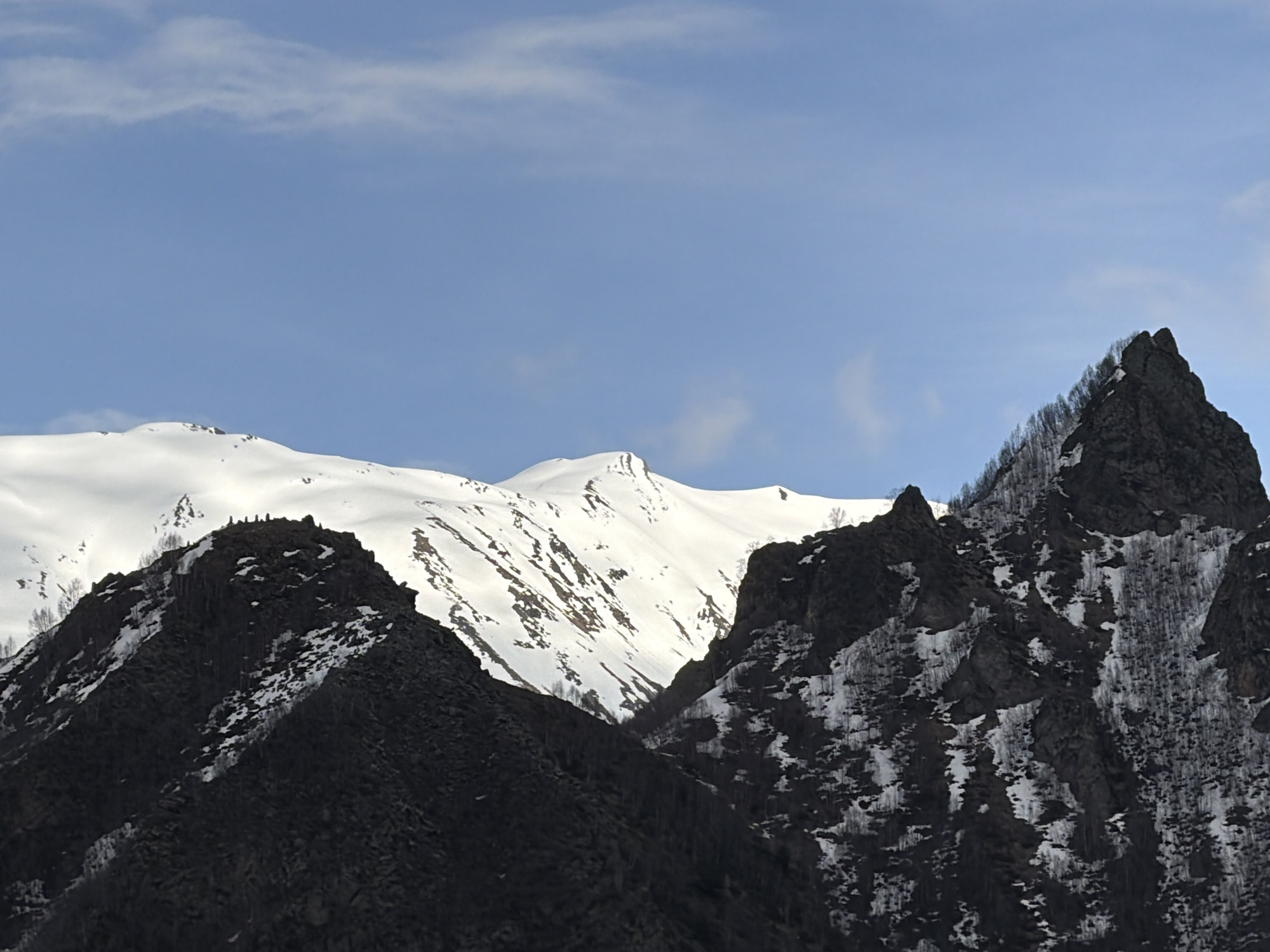 The image shows a mountain landscape in Pkhelshe, Georgia. In the foreground are two dark, rocky mountainsides, one steeply sloped on the left, and a jagged peak on the right. Patches of snow are scattered on the sides of the mountains, and thin trees or bushes are sparsely spread. In the background is a snow-covered mountain range under a blue sky with thin clouds.