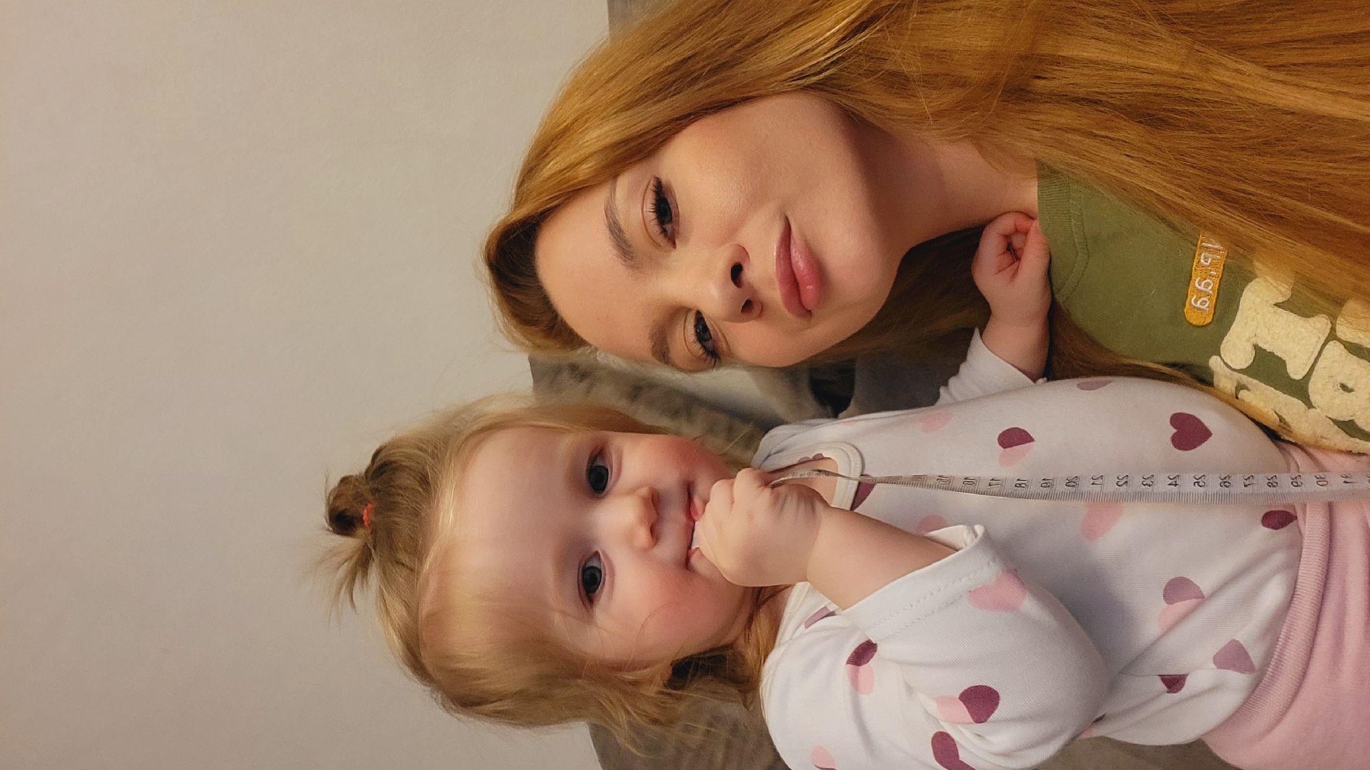 The image depicts a fair-skinned woman with long auburn hair, lying next to a fair-skinned child with blonde hair and rosy cheeks. The child is holding a measuring tape in its hand and appears to be looking directly at the camera. The woman is wearing a green shirt. The child is wearing a white long-sleeved shirt with pink and maroon hearts, and light pink pants. The scene appears to be indoors. There is no information to confirm this image was captured in Chaikovsky, Russia.