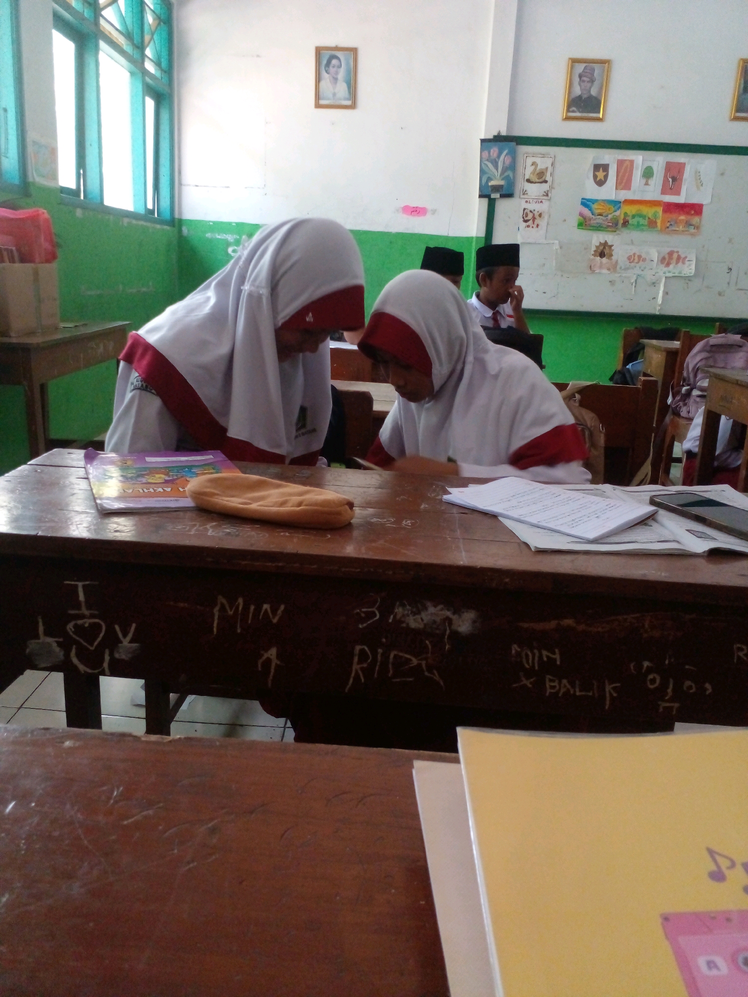 The image captures a classroom scene, likely in Banyuputih, Indonesia, during daylight hours. Two young female students, dressed in white uniforms with red accents and hijabs, are seated at wooden desks, engrossed in their studies. The student on the left is leaning over her desk, while the one on the right is looking down at her work. The desks are old and appear to have graffiti. On the desks, there are books and papers. In the background, a whiteboard is visible, adorned with children's drawings and educational materials. Framed pictures are hanging on the wall. The classroom has green-painted walls below a white upper section, with large windows on the left allowing natural light to enter. The overall impression is one of a typical Indonesian elementary school classroom environment.