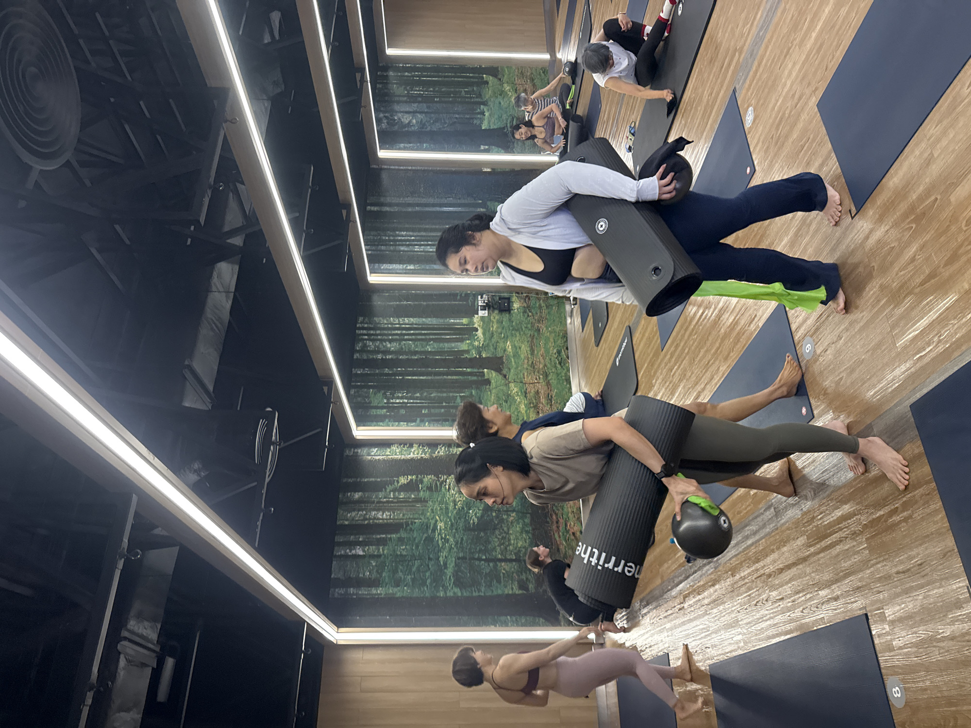 This is a medium shot of a yoga studio in Nong Pa Khrang, Thailand. The studio has a wooden floor and a mural of a forest on the back wall. Several women are practicing yoga in the studio. In the foreground, two women are holding yoga mats. One woman is wearing a grey t-shirt and olive green leggings, and she is carrying a black yoga mat and a small black ball. The other woman is wearing a grey hoodie and dark blue pants, and she is also carrying a black yoga mat. In the background, other women are visible on yoga mats, some are in seated poses, and one is in a kneeling position. The lighting in the studio is bright and even, with prominent LED strip lights running along the ceiling.