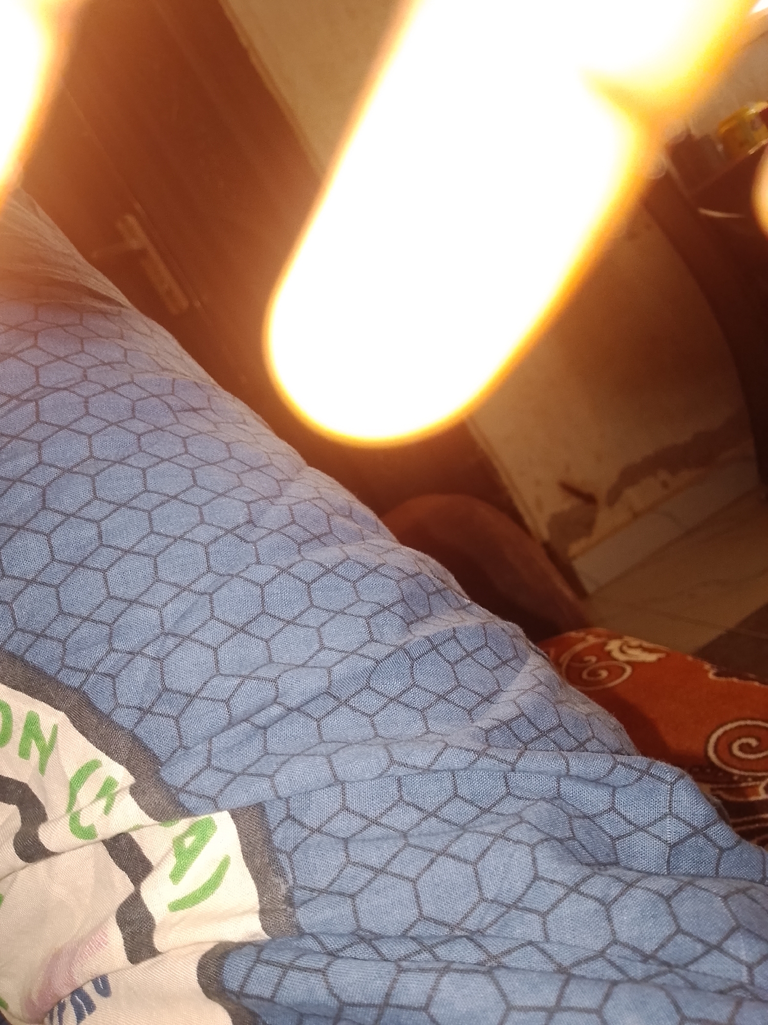 The image shows a close-up view of a person's lap covered by a blue fabric with a hexagonal pattern. The fabric appears to be part of clothing or bedding. There is a bright light source in the upper part of the image, causing a glare and obscuring details. A portion of an orange fabric with a swirling pattern is visible to the right, possibly another piece of bedding or a cushion. The background is dark and out of focus, with hints of a wall and furniture. The scene is likely indoors, possibly in a bedroom. The overall impression is that of a casual, relaxed setting. Without additional context, it is not possible to determine the specific location within Jalingo, Nigeria.