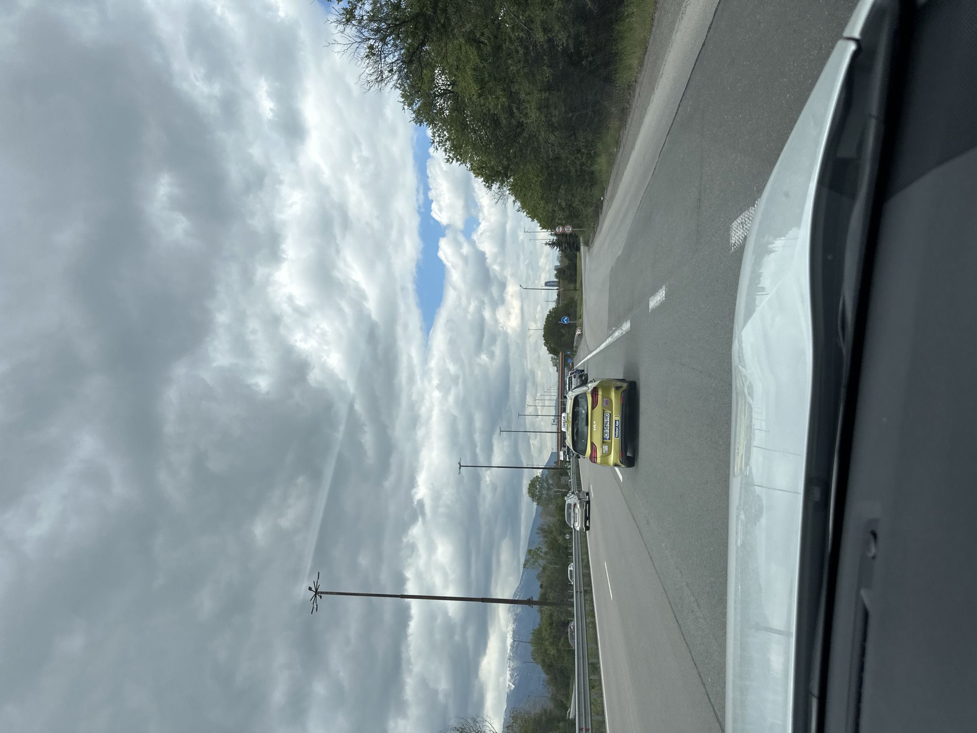 The image shows a view from inside a vehicle moving on a multi-lane highway. A yellow car is traveling in the adjacent lane. Overhead power lines are visible, stretching across the sky. In the distance, mountains are visible beneath a sky filled with gray and white clouds. Based on the license plate visible on the yellow car and the mountain in the distance, the location can be inferred to be in or near Lozen, Bulgaria.
