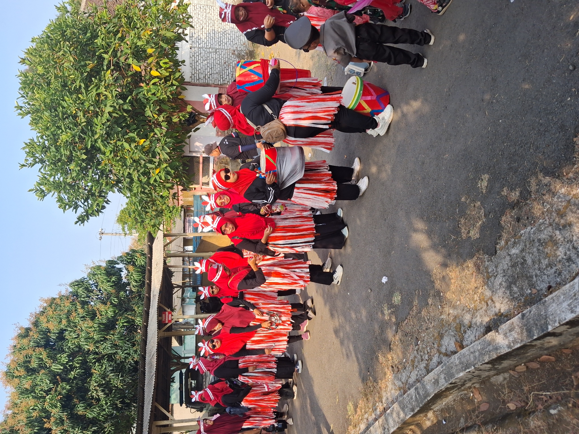In Balapulang, Indonesia, a group of women, dressed in matching red and white outfits and headdresses, are participating in a parade. They are walking on a paved road with a concrete drainage channel on the left side. The women are holding musical instruments, including drums and tambourines, suggesting a musical performance. Some are looking at the camera, while others are engaged with their instruments. In the background, trees and buildings are visible. A young boy in a police uniform is also present, observing the parade. The scene appears to be a community event or celebration.