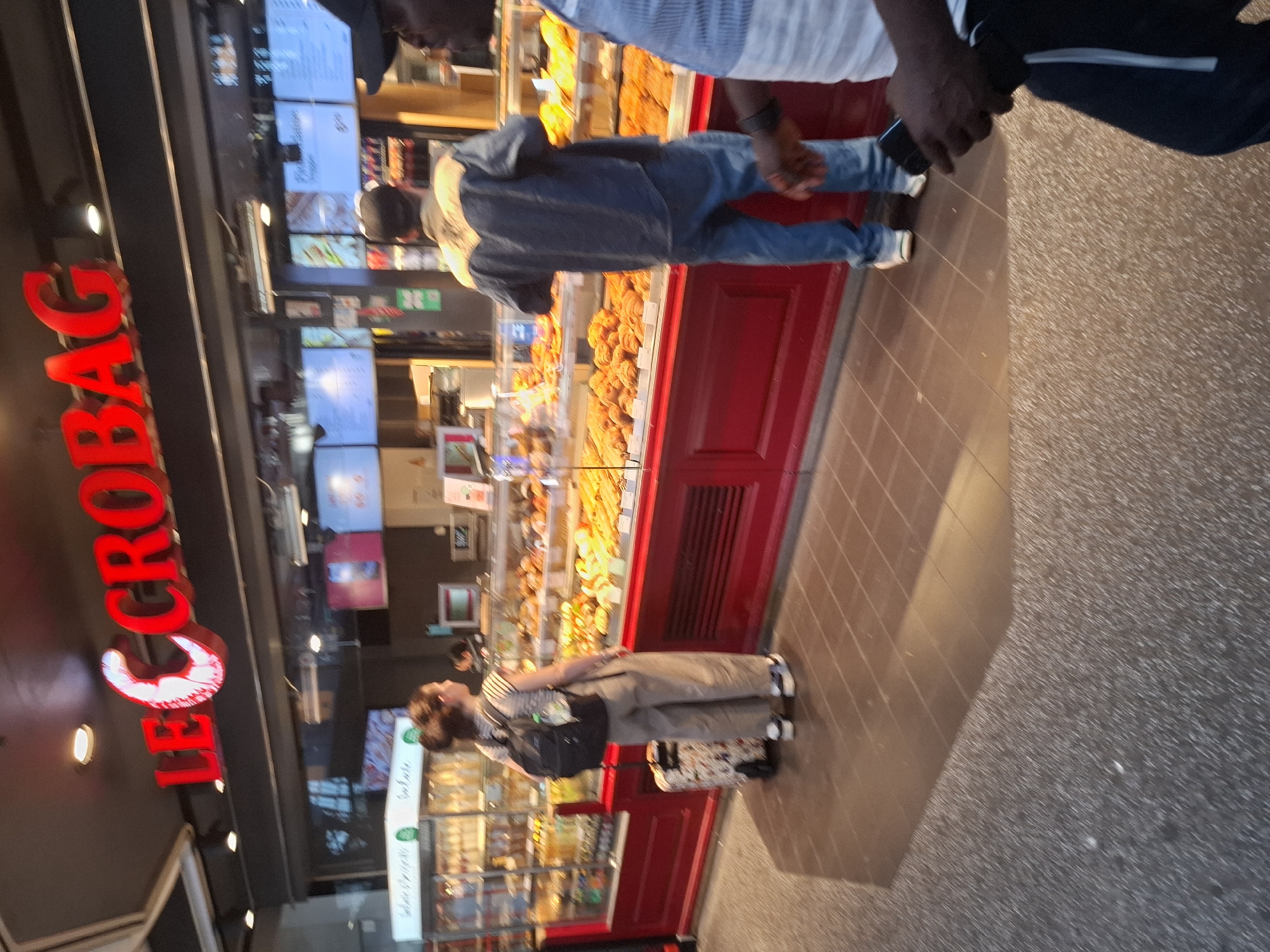 The image depicts the exterior of a bakery in Hamburg, Germany, identified by the prominent red lettering "LE CROBAG" above the shopfront. The bakery appears to be a chain, as suggested by the name. Through the glass display cases, a variety of baked goods are visible, including pastries and breads, arranged on red shelves.
Several individuals are present. A woman with her hair in a bun, wearing a striped t-shirt, beige pants, and a backpack, stands in front of a display case, seemingly contemplating her purchase. Next to her is a suitcase with a distinctive pattern. A man wearing a blue denim jacket and blue jeans stands further down the counter, also looking at the offerings. Partially visible to the right is another person, possibly a customer or an employee, wearing a blue and white striped shirt. The floor is tiled with a reddish-brown pattern. The lighting suggests it is daytime.