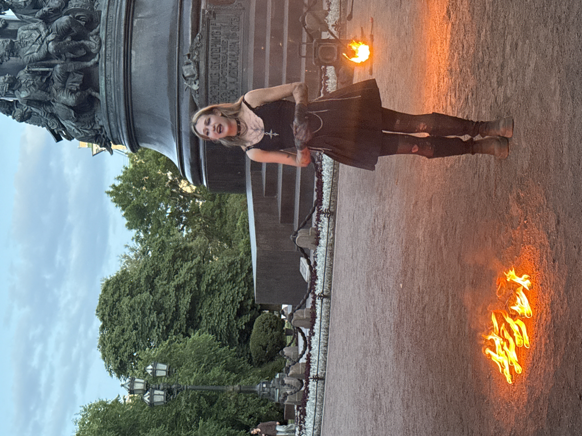 The image captures a scene at the Bronze Horseman monument in St. Petersburg, Russia. A young woman with blonde hair, wearing a black dress, choker, and boots, is standing in front of the monument. She appears to be performing or posing, with her mouth open as if speaking or singing. A small fire burns on the ground in front of her, adding a dramatic element to the scene. The monument itself, a large equestrian statue, dominates the background, with its intricate details and imposing presence. The sky above is cloudy, suggesting it might be late afternoon or evening. Trees and streetlights are visible in the surrounding park area, indicating a public outdoor space.
