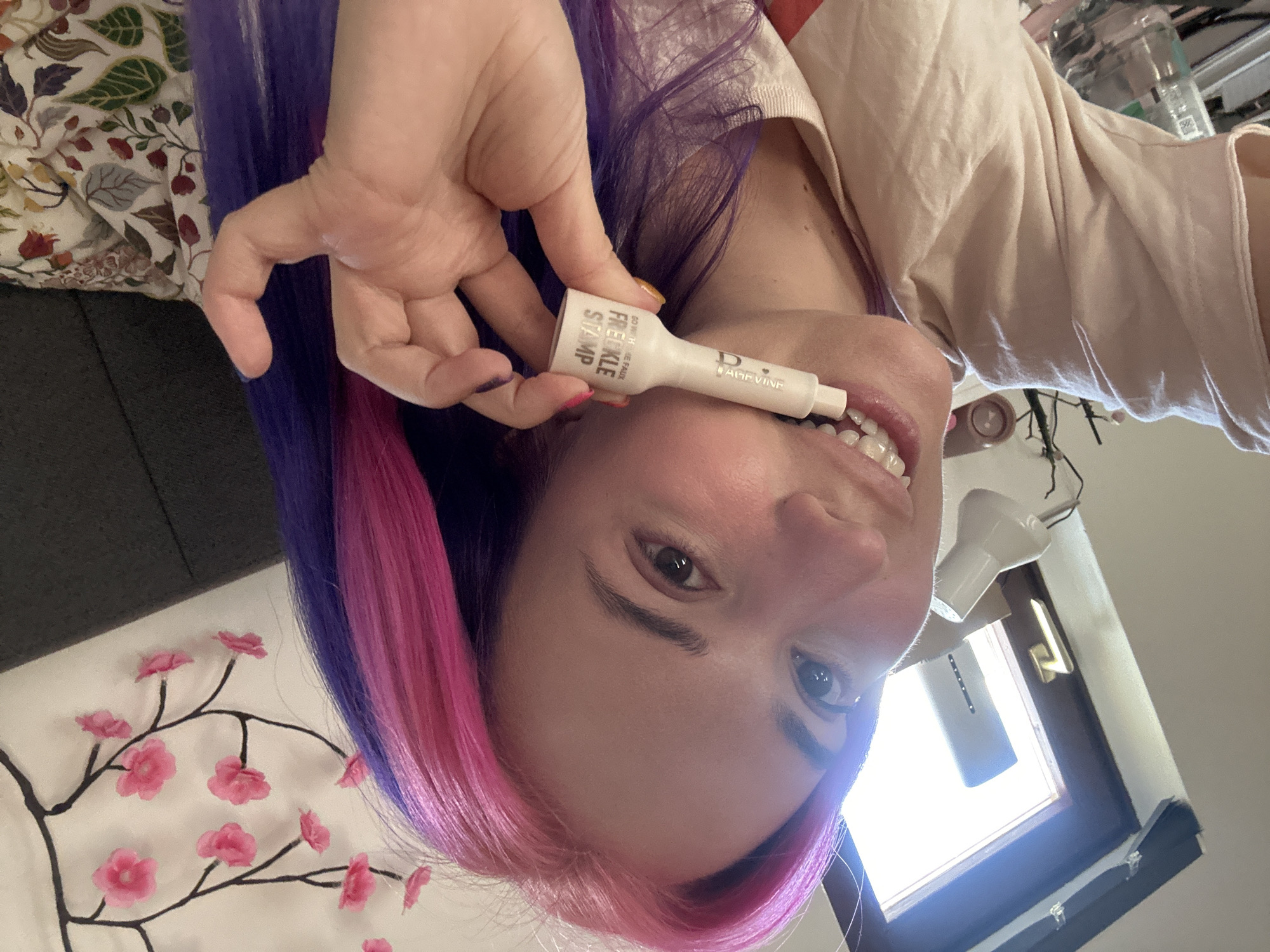 A person with purple and pink hair is holding a white freckle stamp product near their mouth. The background includes a floral patterned blanket, a dark grey object, and a window with natural light. The location is unspecified and there's no clear indication of being in the city of Zagreb, Croatia.