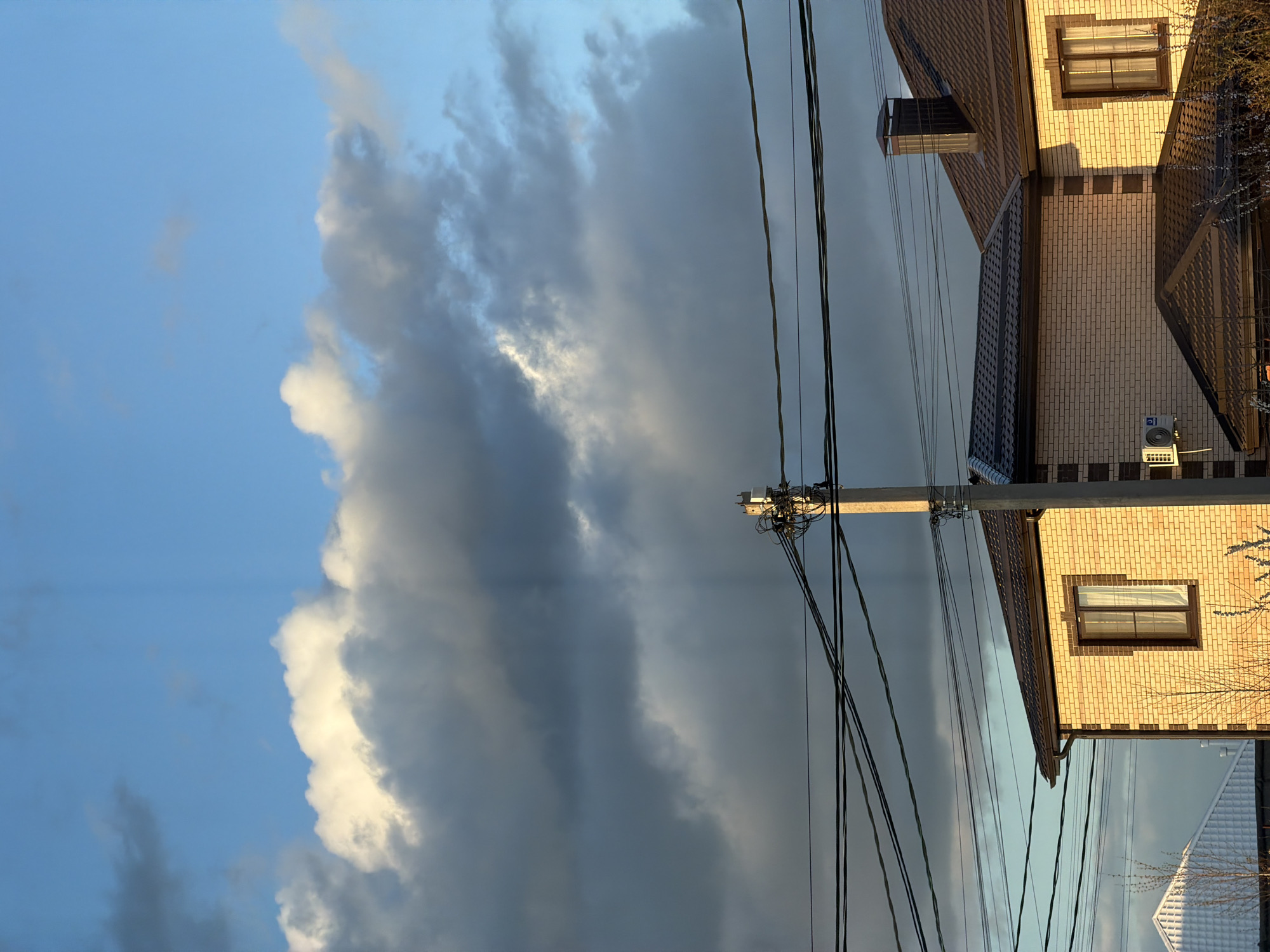 The image depicts a partly cloudy sky above houses and power lines in what appears to be the city of Rossiyskiy, Russia. The clouds are a mix of dark gray and bright white, suggesting a contrast between an impending storm and sunlight. In the foreground, power lines stretch across the scene, connected to a utility pole. To the right of the pole is a two-story house with brick siding, dark roof tiles, and a window with a dark frame. Part of another white house is visible in the bottom corner.