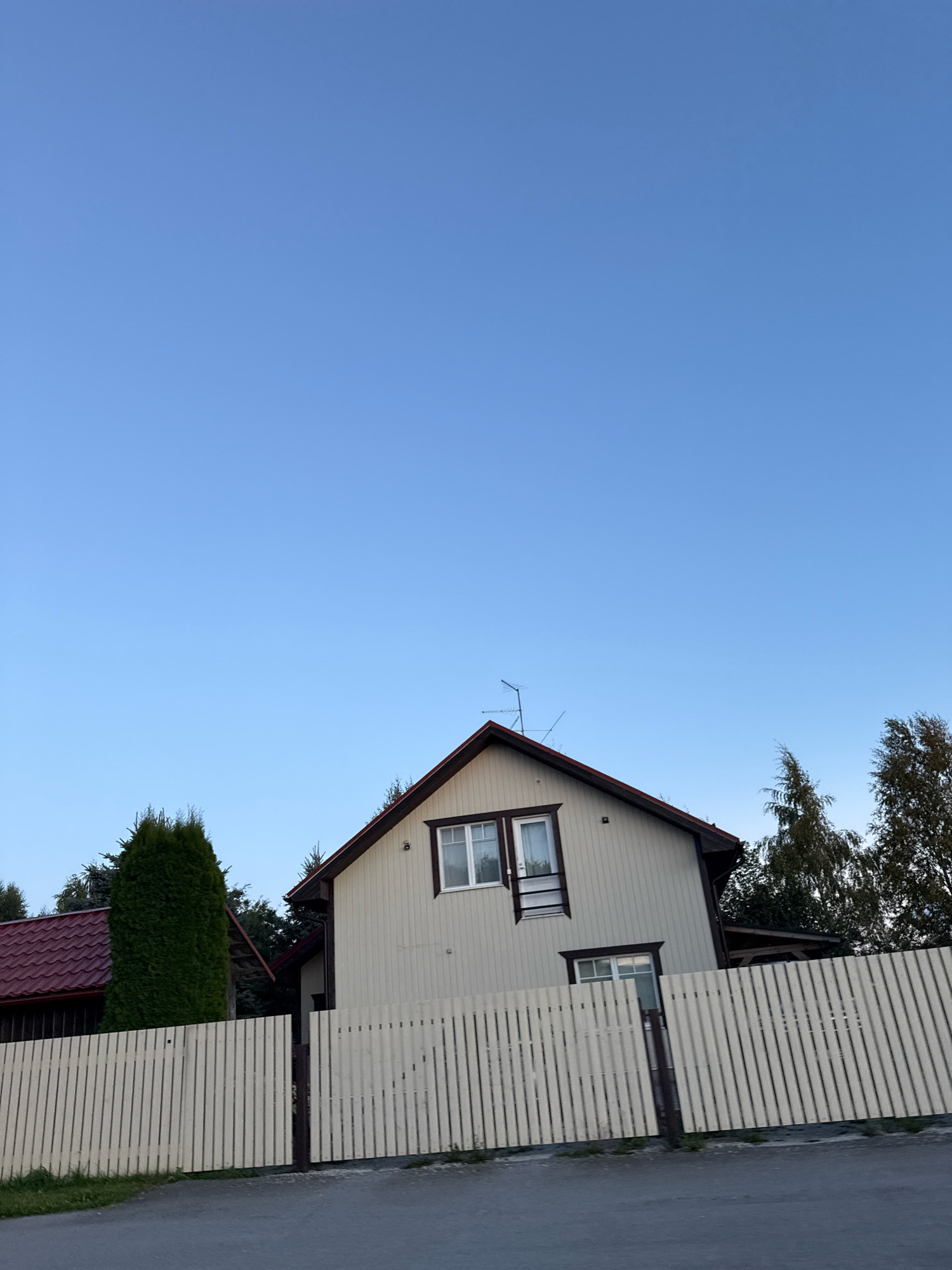 A light-colored, two-story house with a dark red roof is partially visible under a clear blue sky. The house has white-trimmed windows and a television antenna on the roof. A tall, dark green evergreen tree stands to the left of the house, and several deciduous trees with sparse foliage are visible behind the house to the right. A light-colored wooden picket fence runs across the foreground, separating the house from a paved road. The fence is slightly angled and appears to have a gate. The environment suggests a suburban or rural setting in Alliku, Estonia.