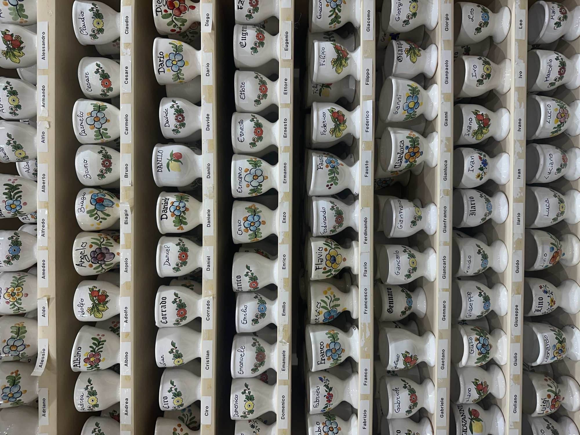 This image presents a detailed view of a wooden display case filled with numerous small, white, ceramic egg cups. Each egg cup is hand-painted with distinct colorful floral designs and features a handwritten Italian male name. Adjacent to each cup, a small white label displays the same name in a clear, printed font.
The names visible on the cups and labels include a wide array of Italian male names such as Alessandro, Alberto, Alfredo, Albino, Aldo, Amedeo, Andrea, Angelo, Armando, Biagio, Bruno, Carmelo, Cesare, Ciro, Claudio, Corrado, Cristian, Daniel, Daniele, Danilo, Dario, Davide, Diego, Domenico, Emilio, Enrico, Enzo, Ermanno, Ernesto, Ettore, Eugenio, Fabio, Fabrizio, Fausto, Federico, Ferdinando, Filippo, Flavia, Franco, Francesco, Gabriele, Gaetano, Gennaro, Giancarlo, Gianfranco, Gianluca, Gianni, Gianpaolo, Giacomo, Giorgio, Giovanni, Giuliano, Giulio, Giuseppe, Guido, Ilario, Ivan, Ivano, Ivo, Leo, Leonardo, Lino, Livio, Luca, Luciano, Lucio, Luigi, Marcello, and Mario.
The organized arrangement within the wooden compartments suggests a retail setting, likely a souvenir or gift shop. The hand-painted nature indicates craftsmanship, offering personalized mementos. The display captures a moment in an indoor commercial space, with no specific activity or people visible, focusing entirely on the array of items. The setting is consistent with a shop found in a tourist location like Taormina, Italy.