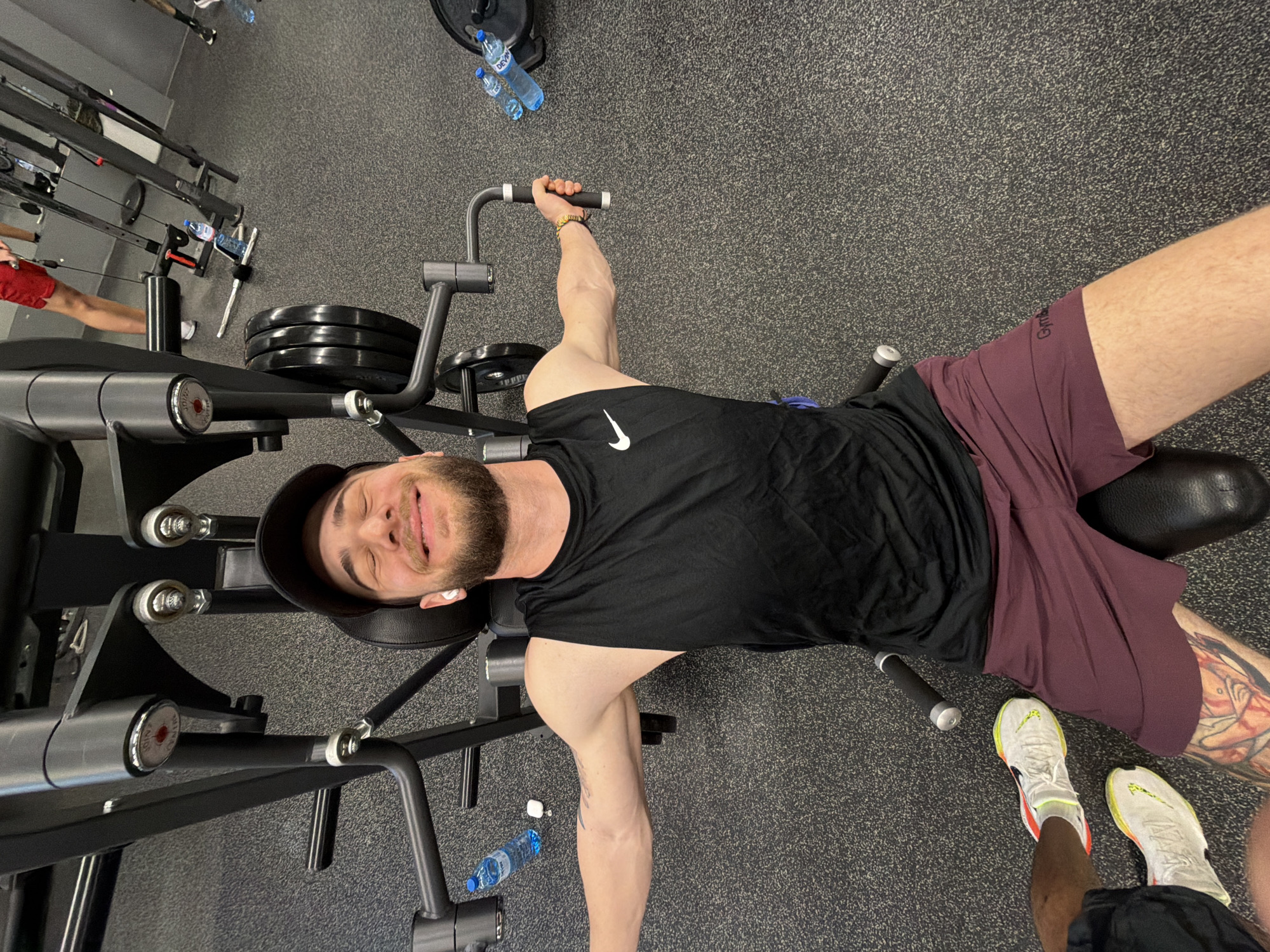 A man wearing a black tank top, maroon shorts, and a black baseball cap lies on a chest press machine in what appears to be a gym. He is smiling at the camera. The gym is gray, with various weight machines and exercise equipment visible in the background. A water bottle is lying on the floor. The image may have been taken in a gym located in Plovdiv, Bulgaria.