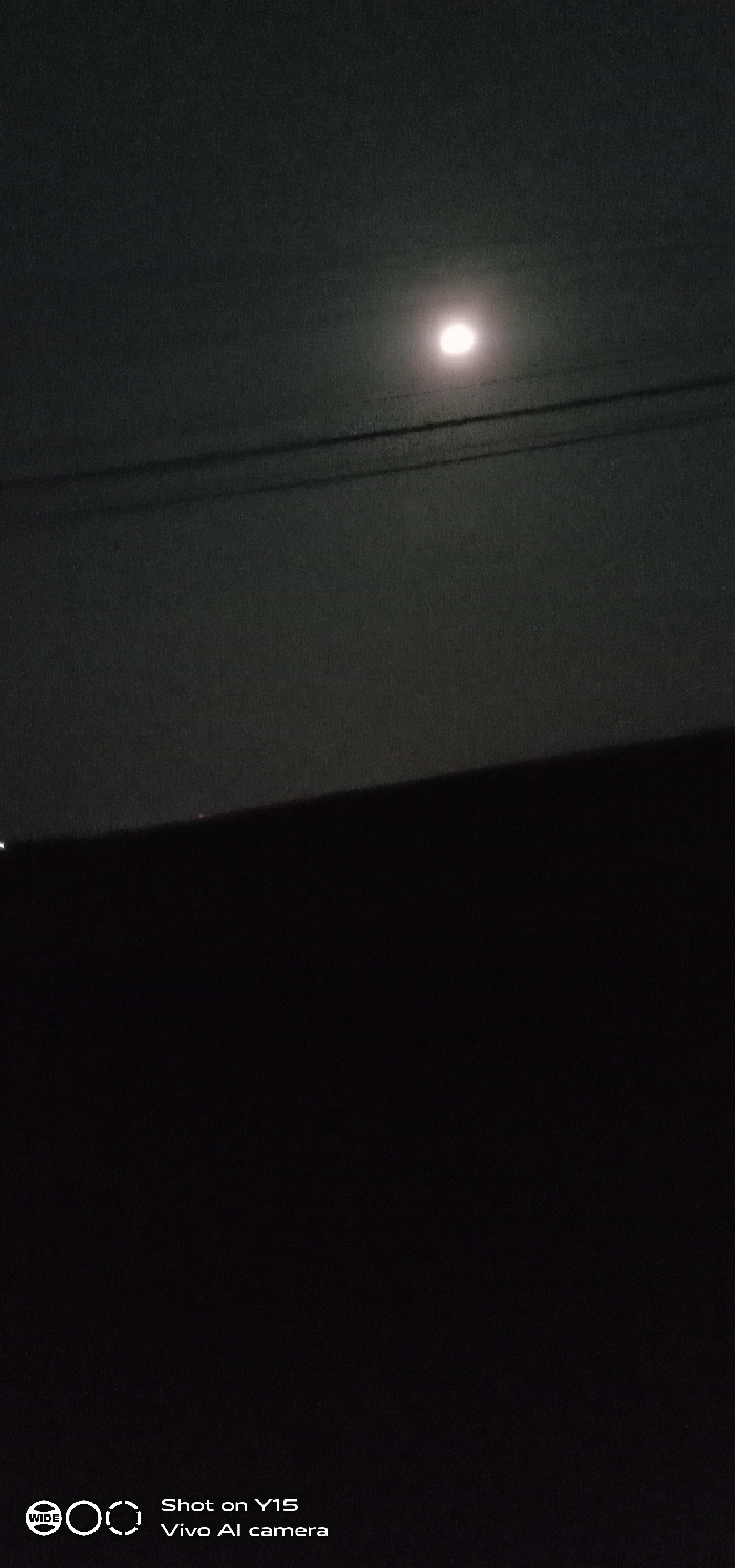 The image displays a night scene captured in Sumberan, Indonesia. The upper two-thirds of the frame show a dark, hazy sky. A single, bright, circular light source, consistent with the moon, is visible in the upper-right quadrant. Below this light source, two distinct, dark, parallel lines, identified as utility wires or cables, extend horizontally across the mid-upper portion of the sky. The lower third of the image is occupied by a solid, dark, horizontal mass, representing a structural element such as a rooftop or wall, which obscures the ground level. A tiny, faint white light is discernible on the far left edge of this dark mass. No people, discernible objects, or specific actions are visible within the primary scene. Text overlaid in the bottom right corner of the image reads "Shot on Y15 Vivo AI camera", and a "WIDE" logo is present in the bottom left corner.