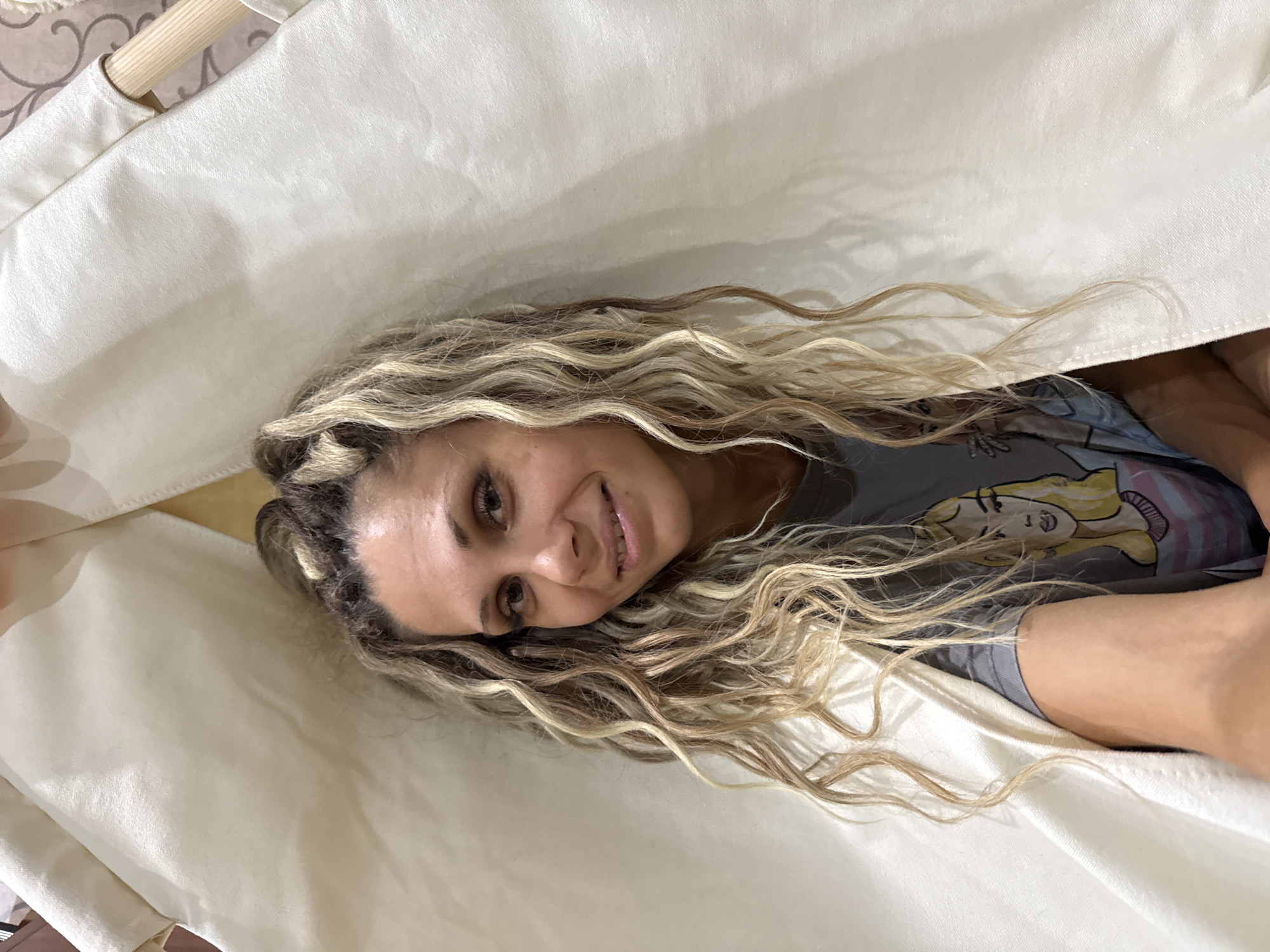 In Rusanovo, Russia, a woman with blonde, curly hair pulled into a bun is pictured from the chest up, looking at the camera with a slight smile. She is wearing a grey t-shirt with a cartoon drawing of a woman on it. The woman is inside what appears to be a teepee or tent made of off-white fabric, which frames her face and upper body. The background is softly blurred, with the teepee’s fabric dominating the image.