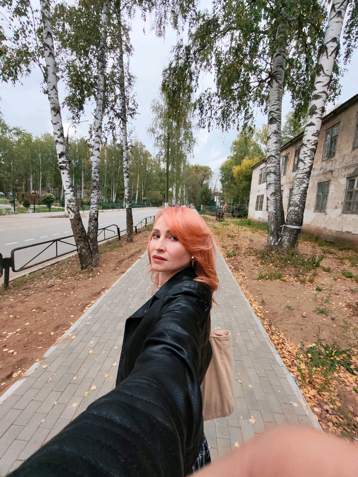 An outdoor, eye-level, medium shot captures a woman with short, peachy-pink hair and light skin, facing the camera from the chest up as she walks along a paved sidewalk. She wears a black leather jacket over a plaid skirt and carries a light brown tote bag on her right shoulder. Her lips are lightly colored, and she wears subtle eye makeup and earrings. The sidewalk is lined with fallen yellow leaves, and a dirt path with more leaves borders the other side of the sidewalk. A dark metal fence runs along the edge of the sidewalk, separating it from a paved road with white markings. Birch trees with distinctive white and black bark line the sidewalk, with their green and yellowing leaves creating a canopy overhead. In the background, an old, weathered apartment building with multiple windows stands next to a line of trees. The sky is overcast and a pale grey. The scene appears to be in the city of Chaikovsky, Russia, given the presence of birch trees and the architectural style of the building.