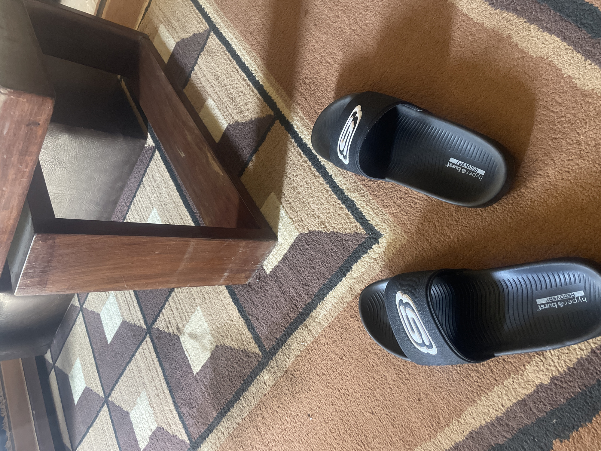 The scene is indoors, showing a brown wooden stool and a pair of black sandals on a brown and tan patterned rug. The sandals are oriented with their toes pointing toward the bottom of the frame. The brand "Hyper Burst" is visible on the sandals. The location is undetermined. The scene lacks any elements that point to it being in Gura Top, Nigeria. No people are visible.
