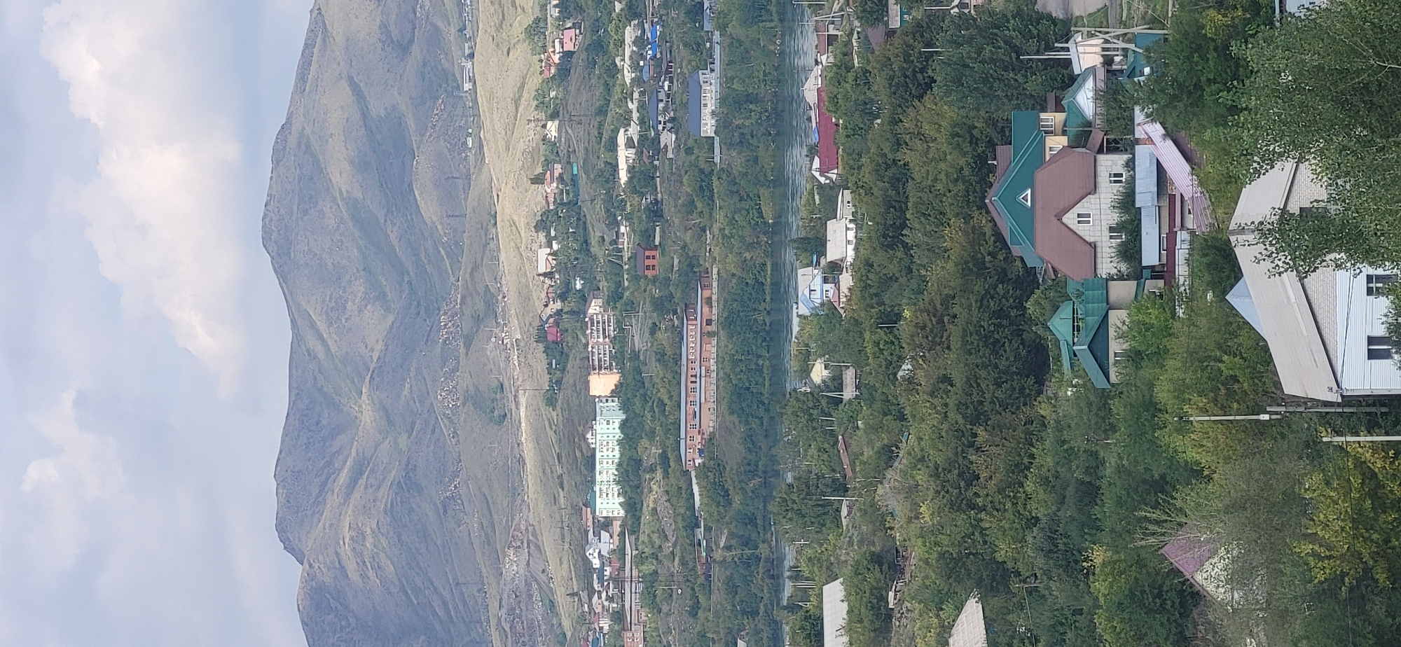 This is an aerial, vertical, high-angle photograph of Oskemen, Kazakhstan, featuring a sprawling residential area nestled at the foot of rolling, arid mountains under a cloudy sky. The middle ground of the image is dominated by a body of water, likely a river, which reflects the overcast sky and is lined by lush green trees. On the far bank of the water, a distinct, long, multi-story building with a brick facade and visible windows stands out. In the foreground and mid-ground, a dense collection of houses of various sizes and architectural styles are interspersed with abundant green trees and foliage. The houses feature diverse roof colors, including red, brown, green, and grey. Some buildings are single-family residences, while others appear to be apartment complexes. The overall impression is of a settled community harmoniously integrated with its natural mountainous landscape. The image captures a typical daytime scene with no visible human activity or vehicles.