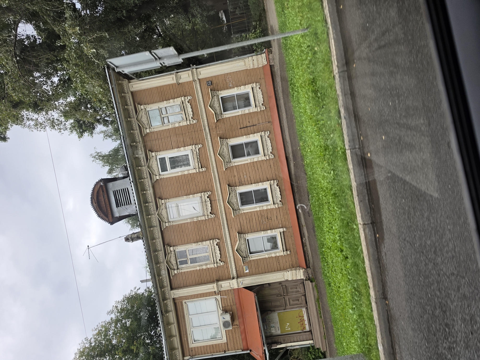 The image captures a tilted view of a two-story, wooden house with a brown facade and white decorative trim around the windows and roofline. The house has a red awning over the entrance on the left side, which features two doors, one of which is yellow and covered in graffiti. An air conditioning unit is mounted to the wall above the awning. The upper floor has multiple windows, each adorned with intricate white carvings. A distinctive dormer window with louvers is visible on the roof, topped with a wooden, curved structure. An antenna extends from the roof. The house is situated on a street with a grassy area separating it from the road, which shows signs of recent rain with puddles visible. Tall trees with green foliage are present in the background, partially obscuring the view of the sky, which is overcast. The number "15" is visible on the side of the house. The overall impression is that of a residential street in the city of Nizhny Novgorod, Russia, likely in a historic district given the architectural style of the house. There are no people or animals visible in the image, and no specific actions are taking place.