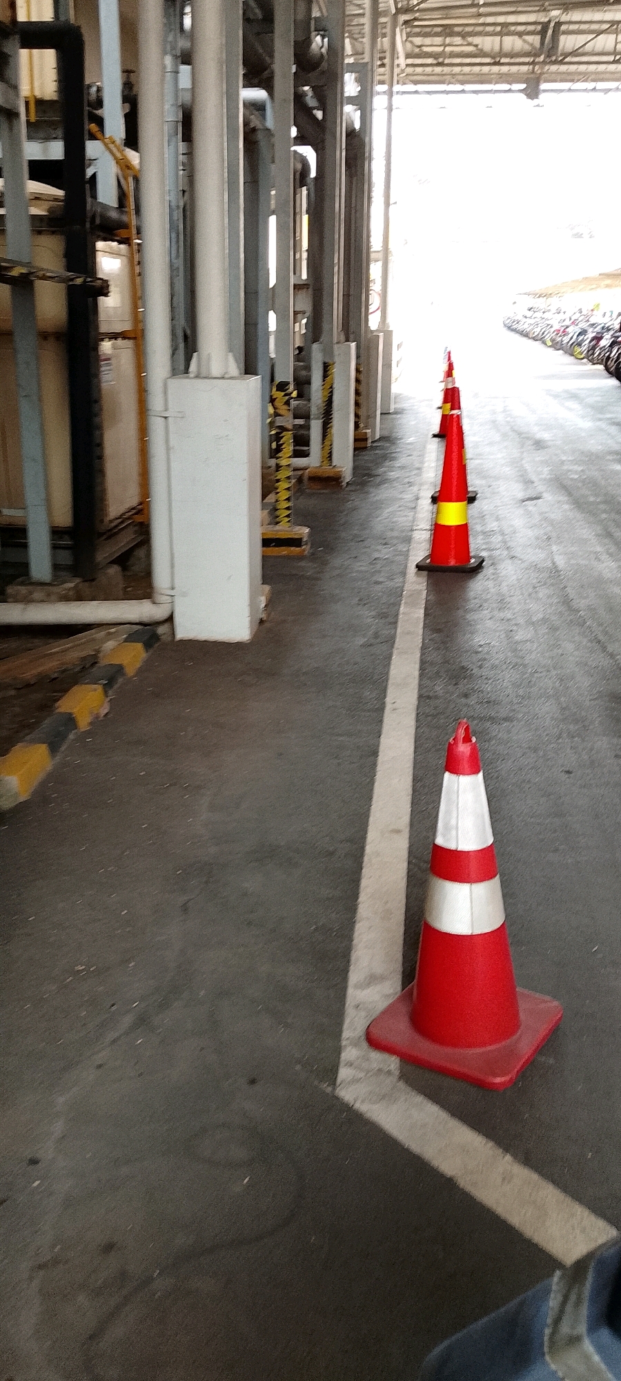 The image depicts an outdoor scene, likely an industrial area or parking lot, with a row of red and yellow traffic cones lining a white painted line on asphalt. In the background, a bright, overexposed area suggests an open space or exit, possibly leading to a road. To the left, there are industrial structures with pipes and metal supports. A line of what appear to be motorcycles or scooters is visible in the distance, parked along the right side of the frame. The scene is captured from a low angle, focusing on the cones and the ground. The location is identified as Depok, Indonesia.