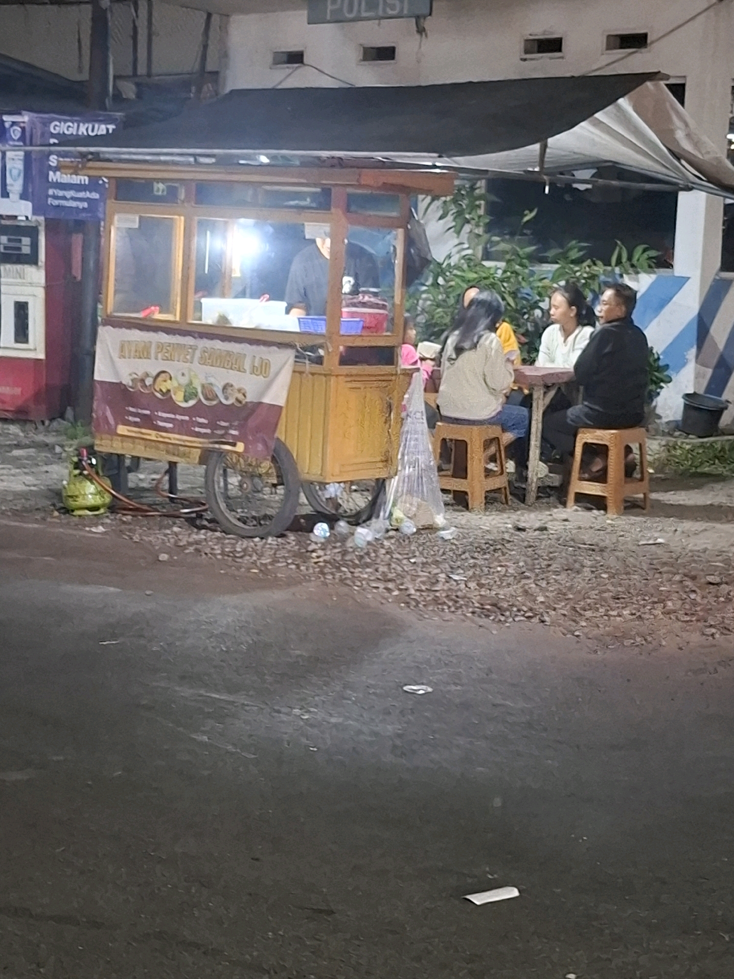 The image depicts an outdoor scene at night, likely in Muka, Indonesia, featuring a food cart and several individuals. A yellow food cart with a banner displaying Indonesian text and images of food items is prominent. The banner reads "AYAM PENYET SAMBAL IJO". A propane tank is visible next to the cart. Three people are seated at a table in front of the cart, facing away from the camera. A fourth person, a woman in a light-colored top, is partially visible at the left side of the table. The individuals are seated on small, orange stools. In the background, a building with a white facade and horizontal blue stripes is visible. There is also signage on a dark blue background with white text above the food cart, indicating "GIGI KUAT" and "Malam". The ground is covered in gravel and debris. The overall impression is a casual, late-night dining setting.